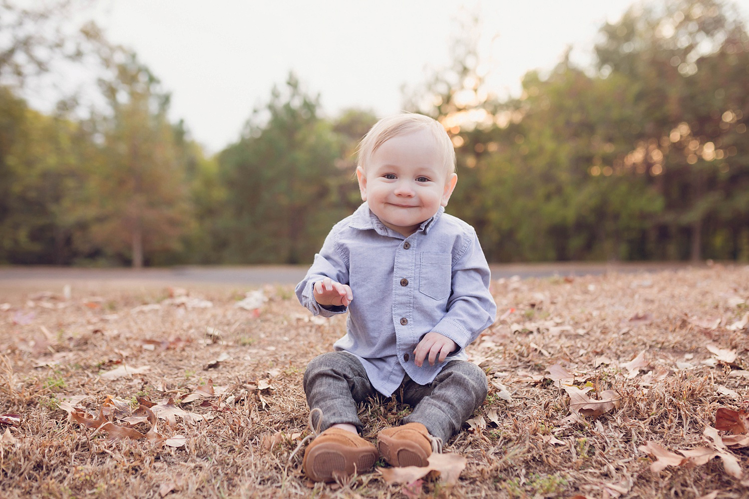  A happy little baby dressed in a blue oxford shirt and brown linen pants with suede shoes sits on the ground and smiles at the camera while laughing in Atlanta.&nbsp; 