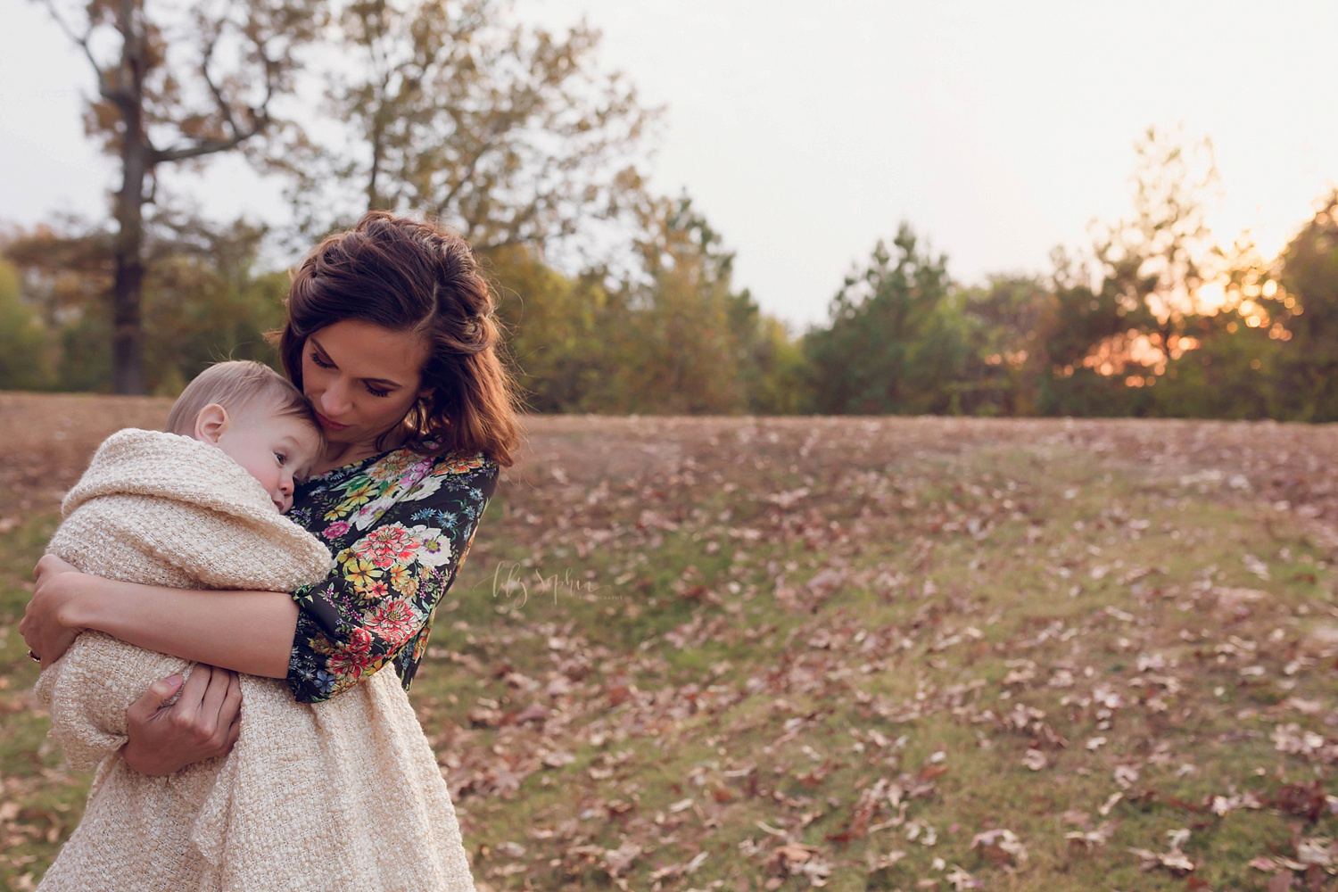  A mother in a floral maxi dress holds her baby son wrapped in a blanket with the sunset sky behind her in an Atlanta park.&nbsp; 