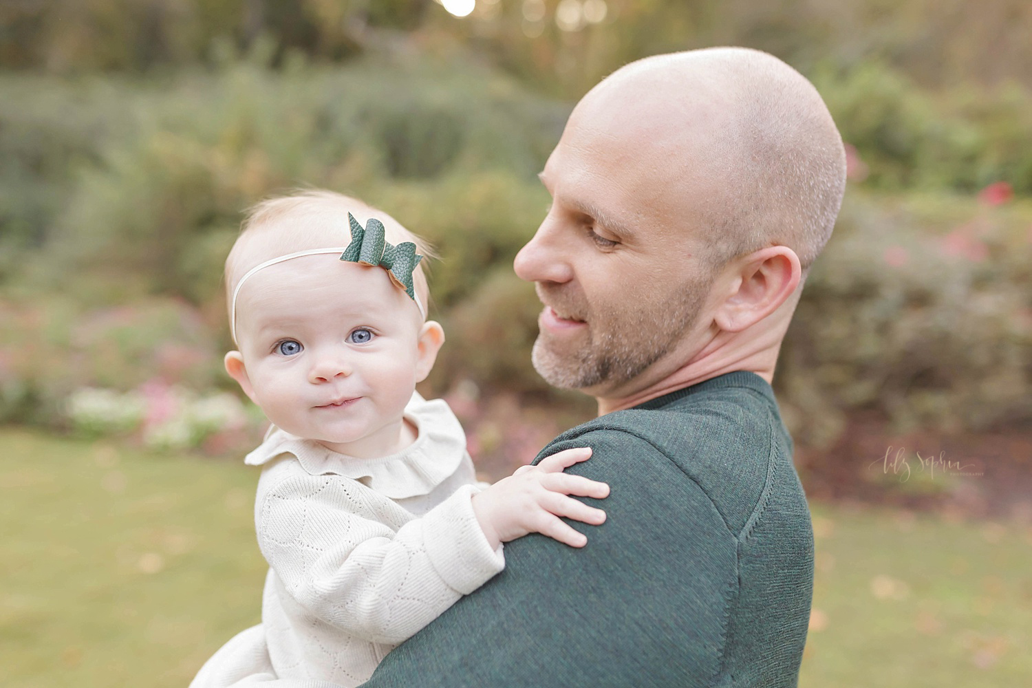  Image of a baby girl wearing a white dress, with a green, polka dot bow headband, being held by her smiling father.&nbsp; 