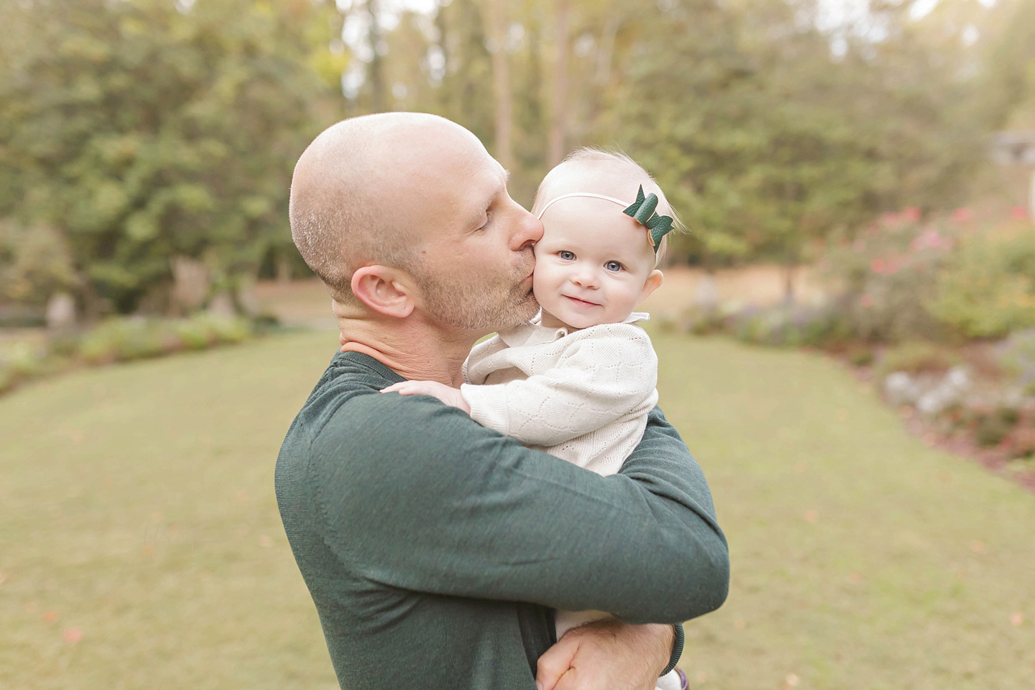  Image of a baby girl wearing a white dress and a green polka dot bow headband while being held and kissed on the cheek by her father.&nbsp; 