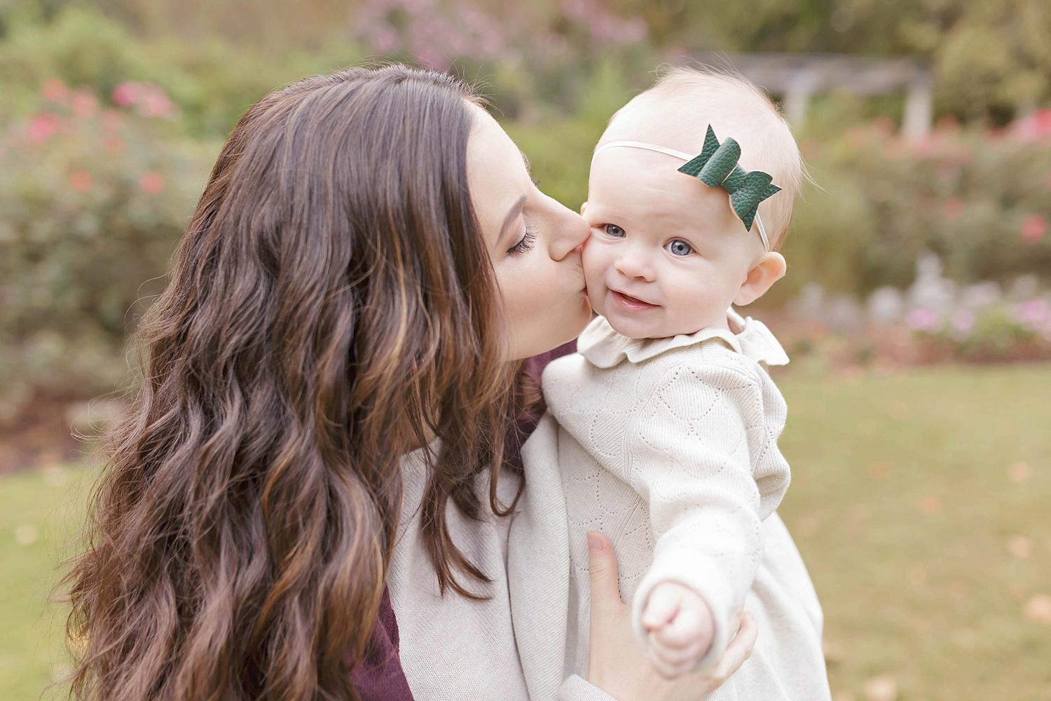  Image of a baby girl wearing a white dress and a green polka dot bow headband, getting kissed on the cheek by her mother. 