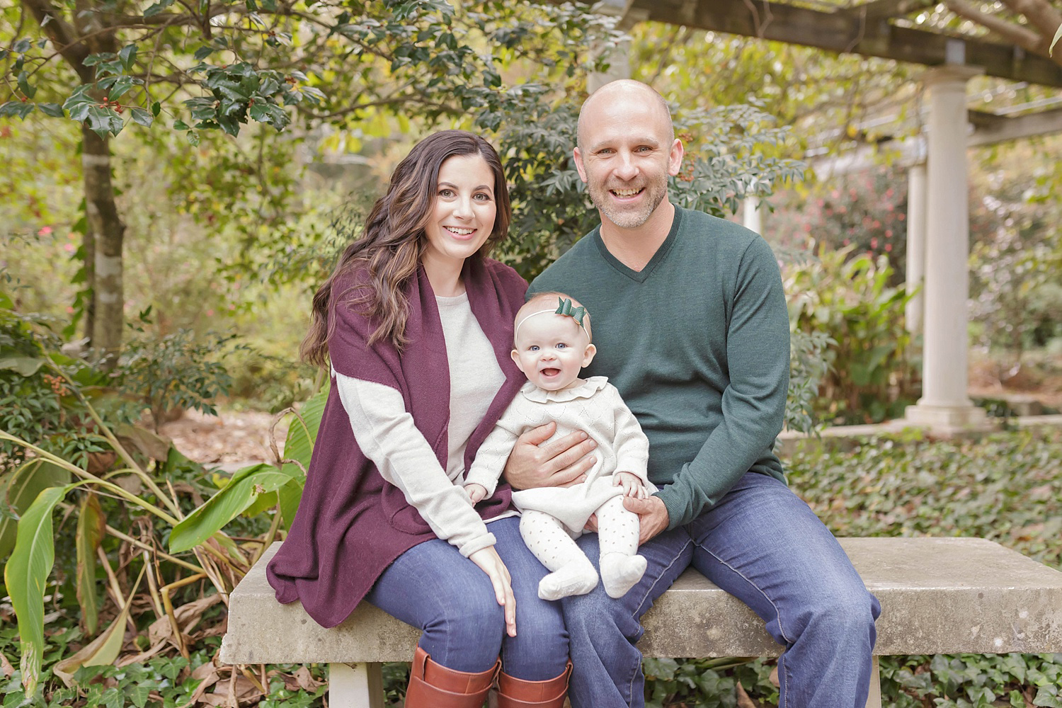  Image of a family of three sitting on a stone bench smiling at the camera. Taken in the garden location of Lily Sophia Photography.&nbsp; 