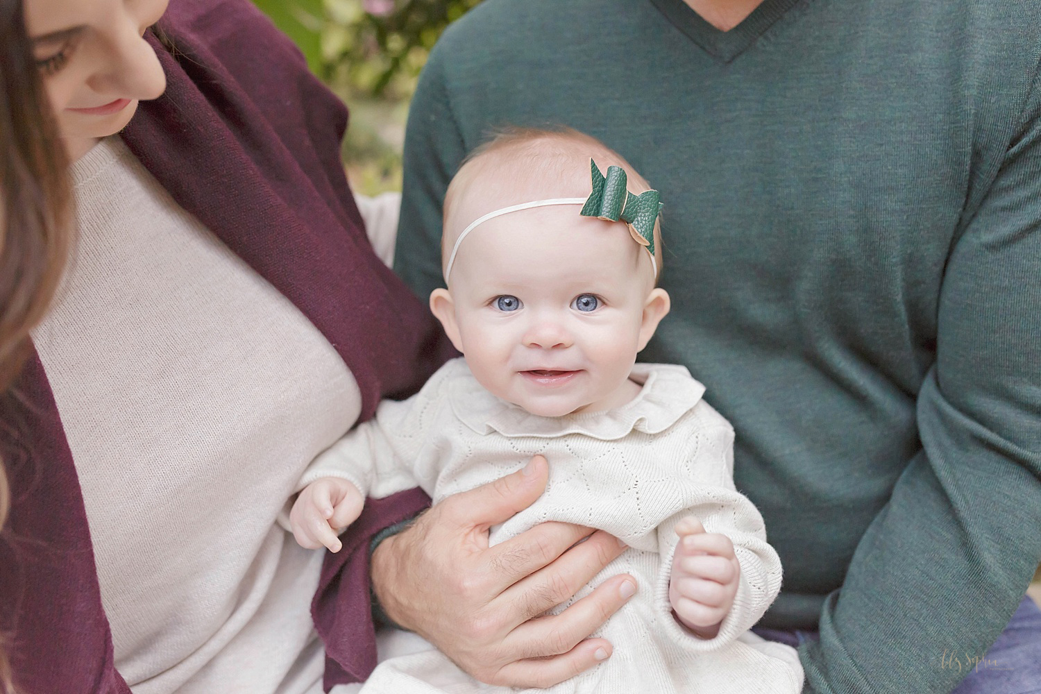  Image of a smiling,&nbsp;blue eyed, baby girl wearing a white dress with a ruffled collar and a green polka dot headband, being held by her parents. 