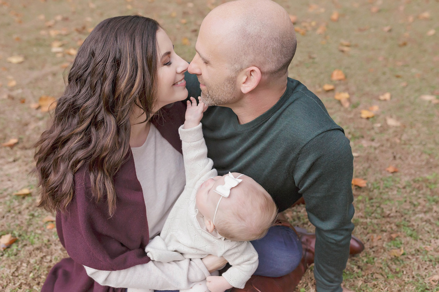    Image of a mom and dad touching noses while holding their baby girl who is reaching up trying to touch their faces. 