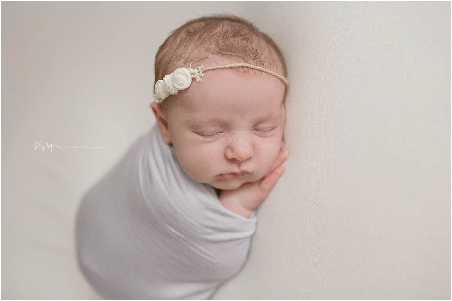 Image of sleeping newborn baby girl in Atlanta natural light studio wrapped in lavender wrap with her hand under her cheek and a white rosebud flower headband on her head.