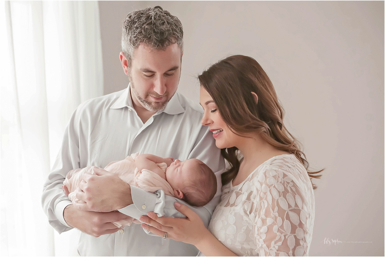 A portrait of a father holding his newborn daughter while the mother touches his arm as they both smile down at their newborn daughter.