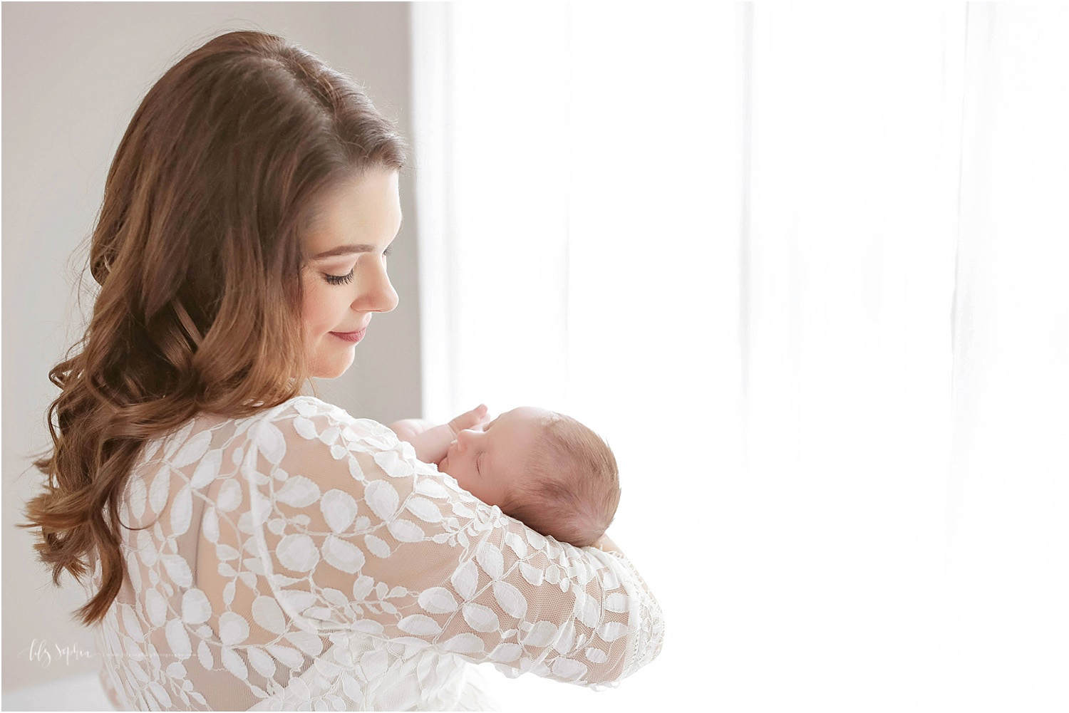 A mother in a white lace dress, smiling and looking down at her newborn daughter in her arms taken at a natural light studio in the Grant Park neighborhood of Atlanta.&nbsp;