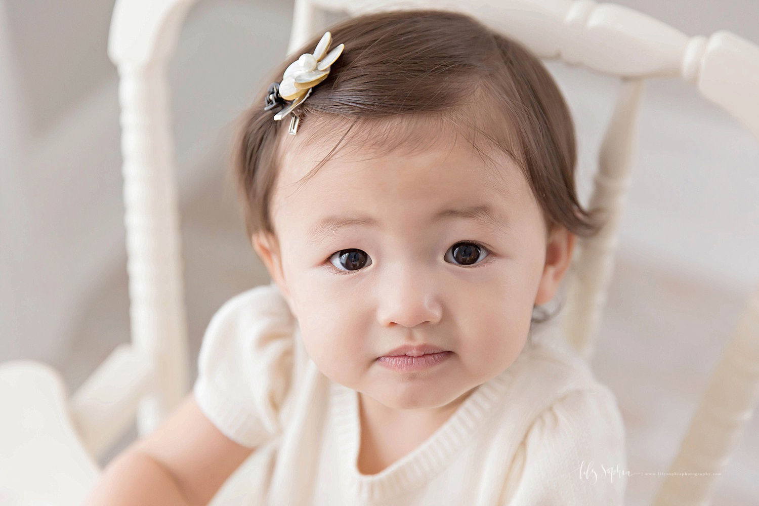  Image of an Asian, baby, girl wearing a cream shirt with a flower clip in her hair, sitting in an antique wooden high chair, and looking up at the camera.&nbsp; 