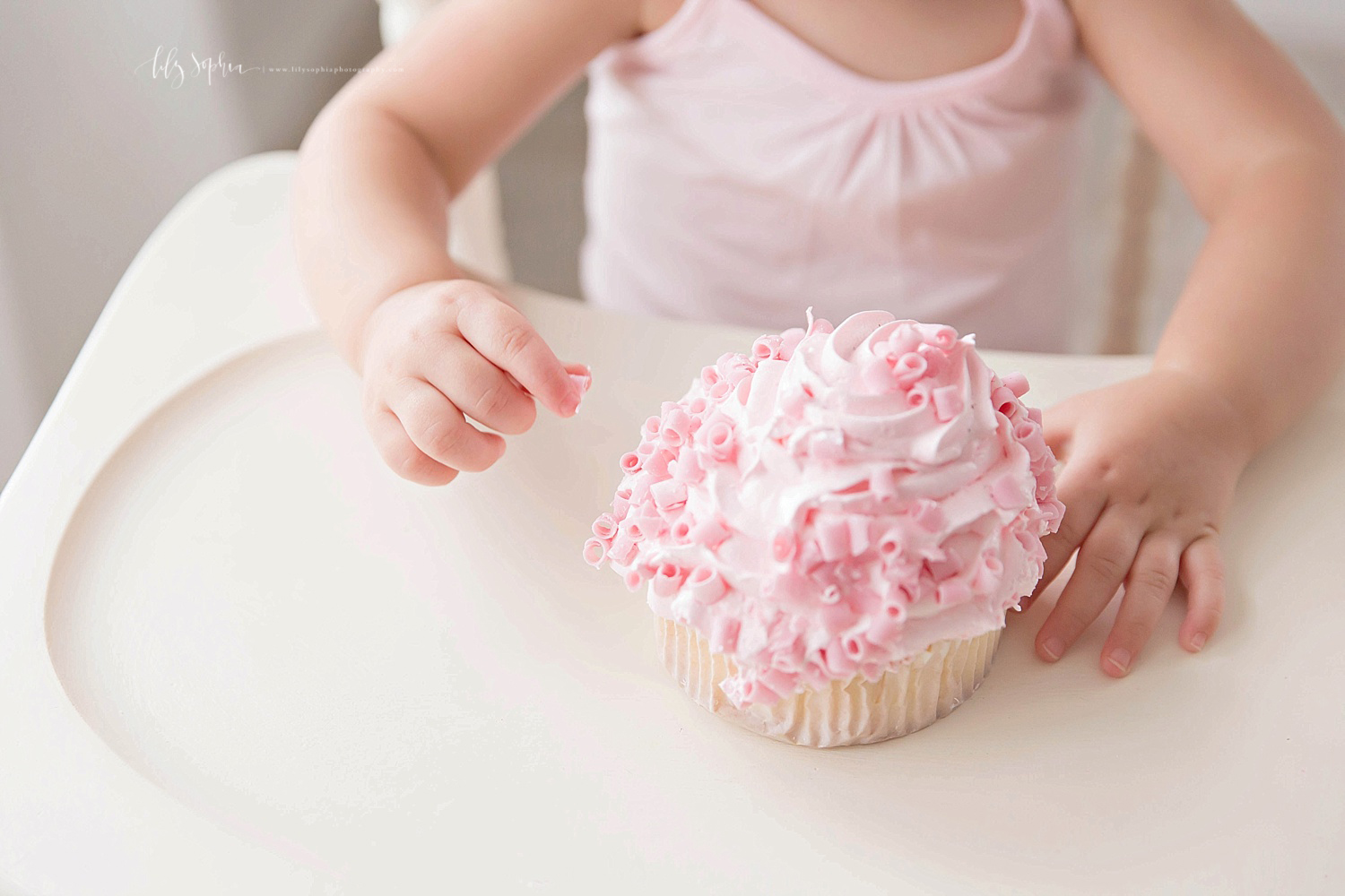  Close up Image of a pink, ruffled, cupcake on a wooden high chair tray, with little hands read to dig in. 