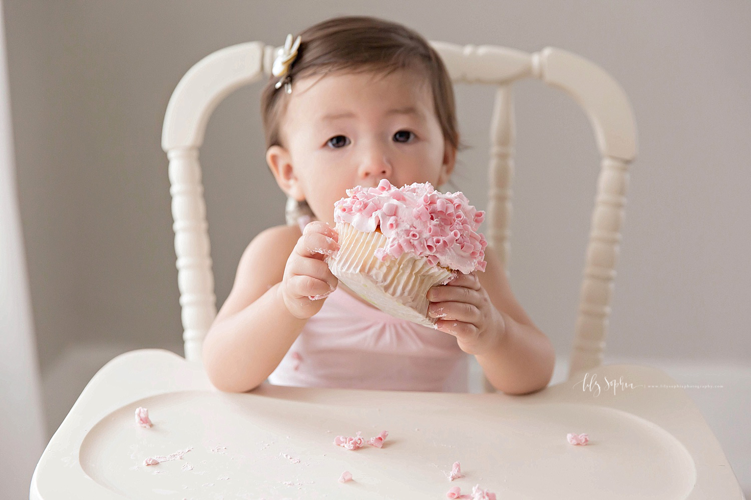 Image of an Asian, baby, girl, picking up her entire pink cupcake, and putting it in her mouth.&nbsp; 