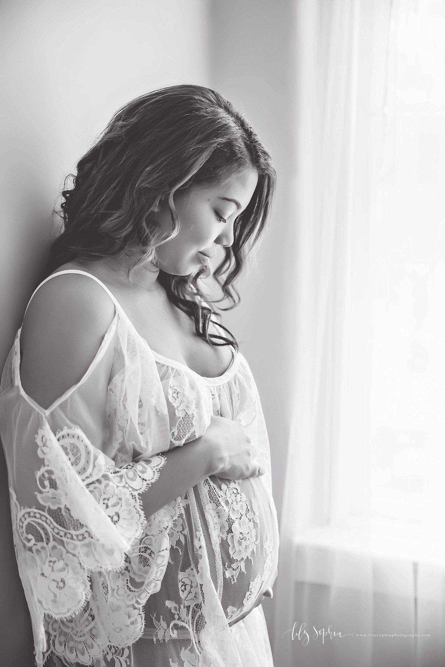  Black and white image of a pregnant, African American, woman, wearing a cold shoulder, sheer lace dress, and holding her belly.&nbsp; 