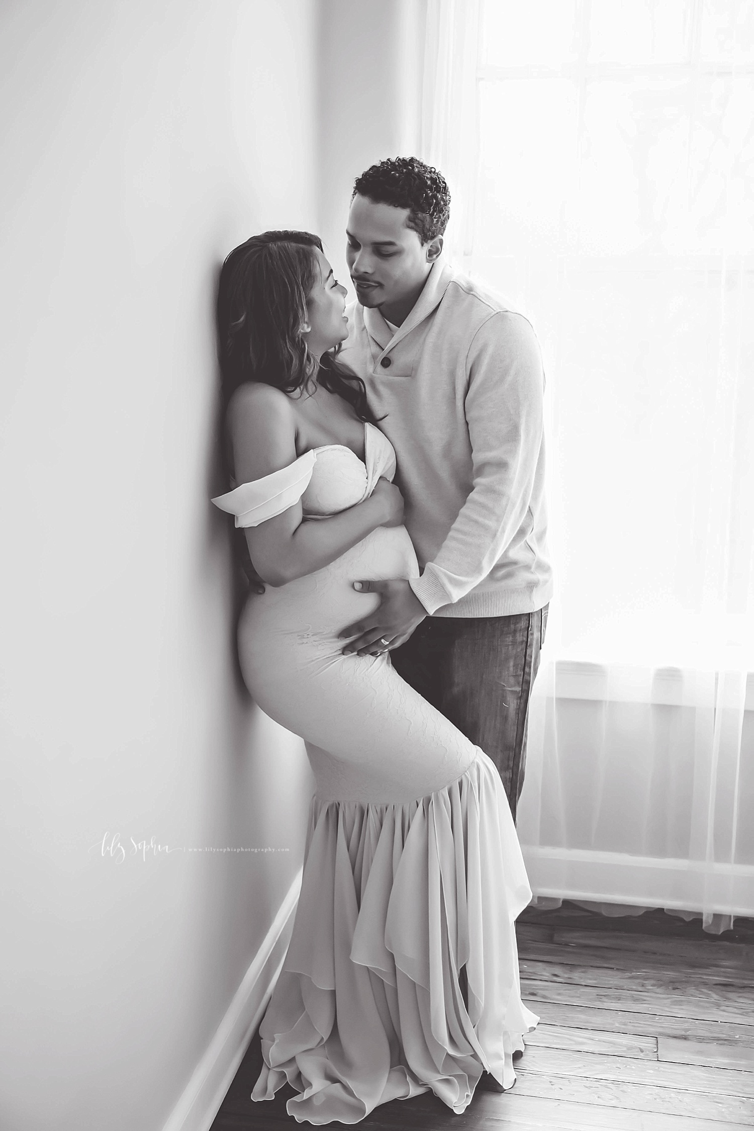  Image of a pregnant, African American woman leaning against a wall, wearing an off the shoulder, mermaid dress, and looking at her husband, smiling while he holds her, with one hand on her belly.&nbsp; 