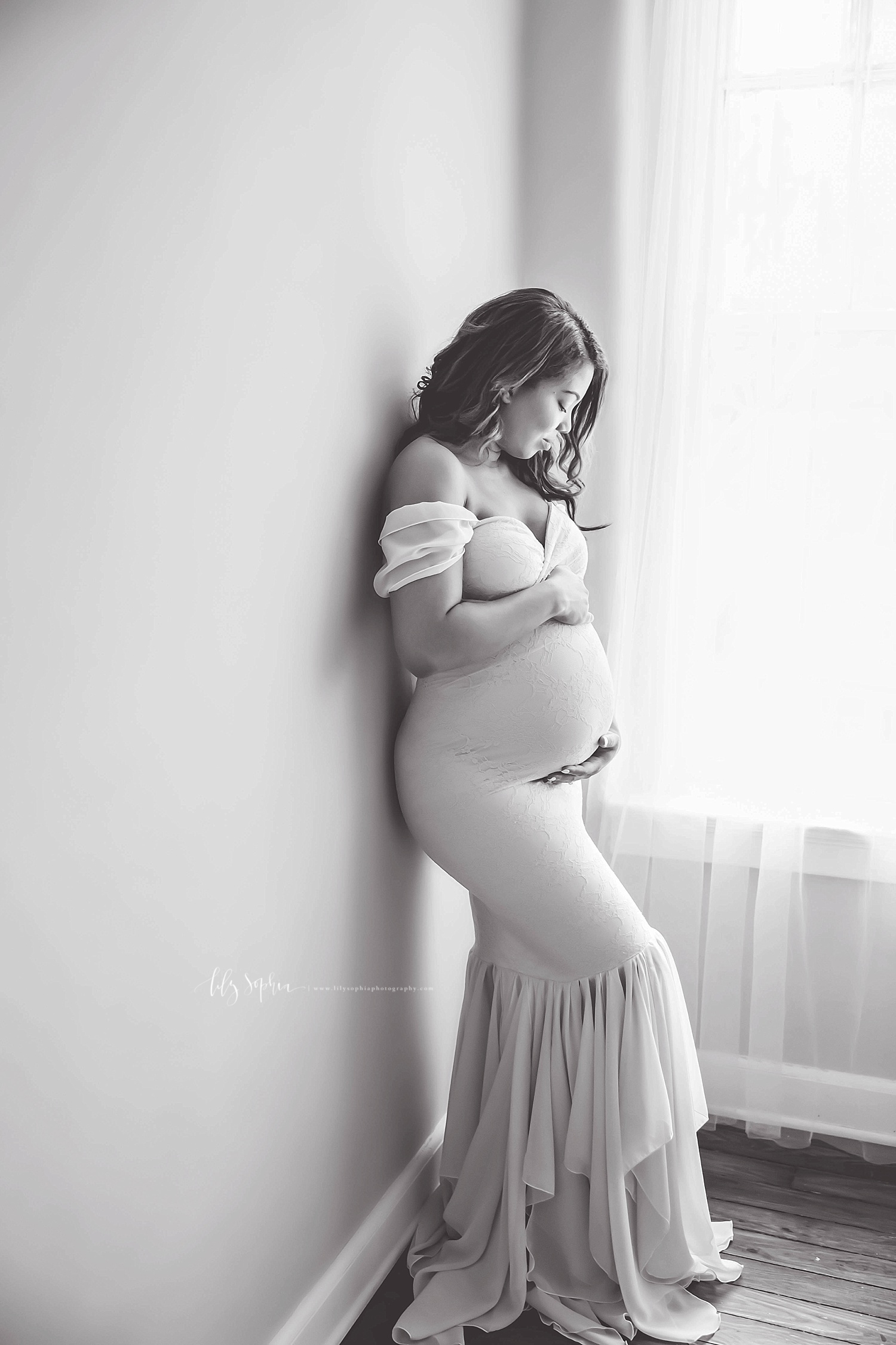  Black and white image of a pregnant, African American, woman, wearing an off the shoulder mermaid dress, leaning against a wall and holding her belly.&nbsp; 