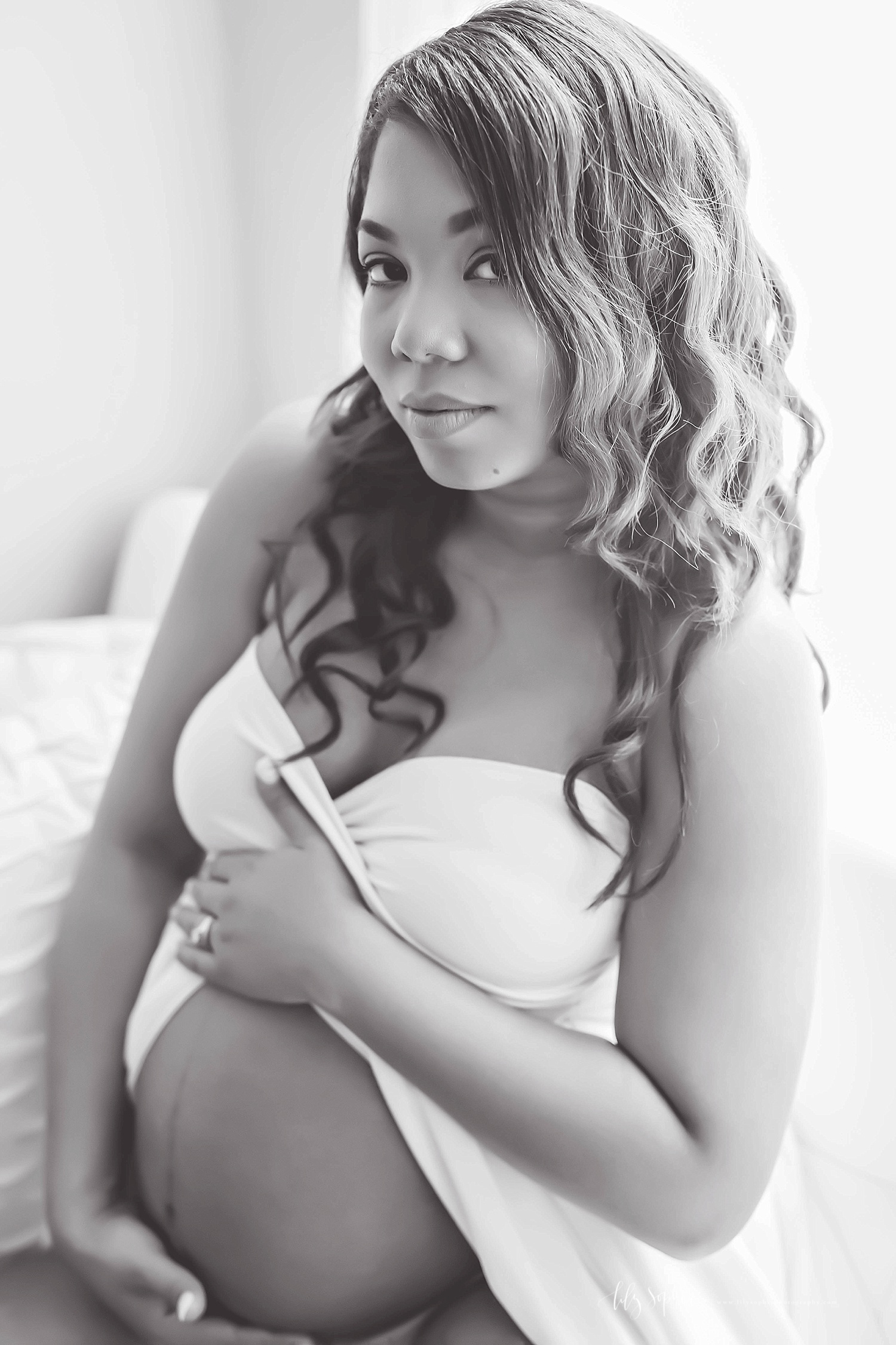  Black and white image of a pregnant, African American woman, sitting on a bed, wearing a strapless, split front, dress, holding her bare belly.&nbsp; 