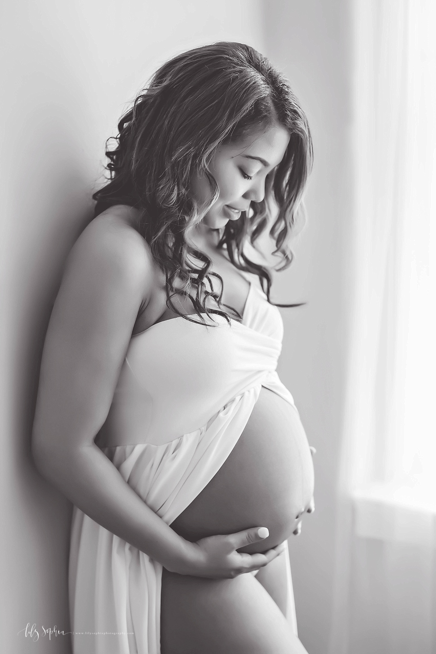 Black and white image of a pregnant,&nbsp;African American, woman, leaning against a wall, wearing a strapless, split front, dress, looking down, and holding her bare belly. 