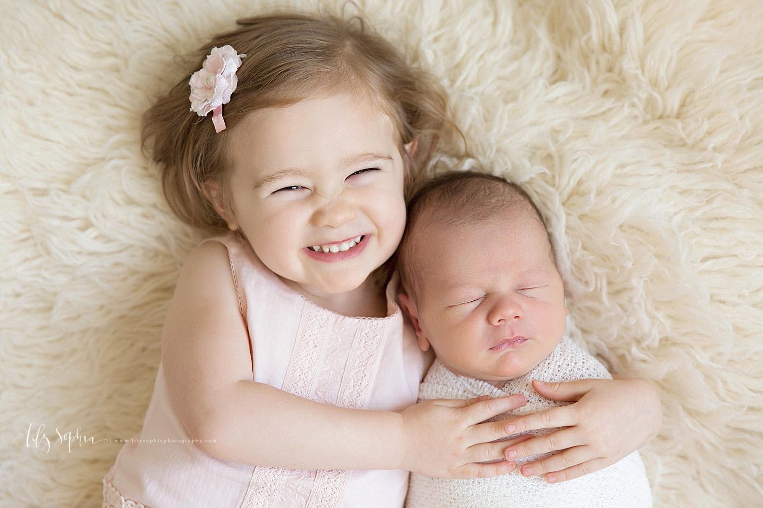  Image of a toddler girl, holding laying down on a cream flokati and holding her sleeping, newborn, baby, brother. 