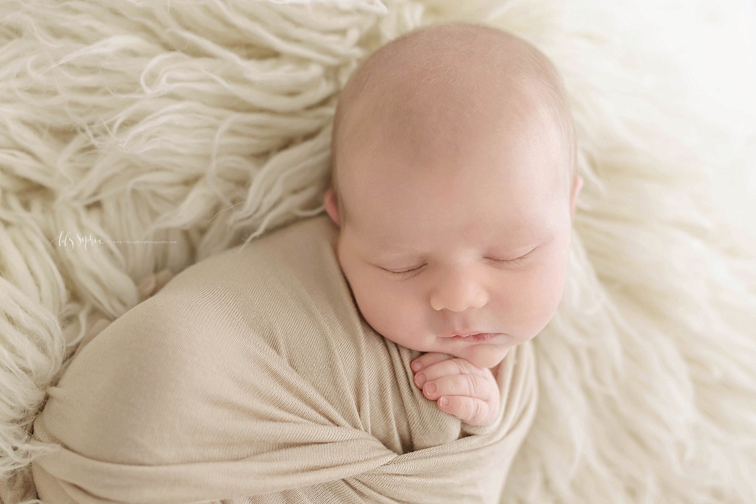  Image of a sleeping, newborn, boy, wrapped in a tan blanket and laying on a cream flokati. 