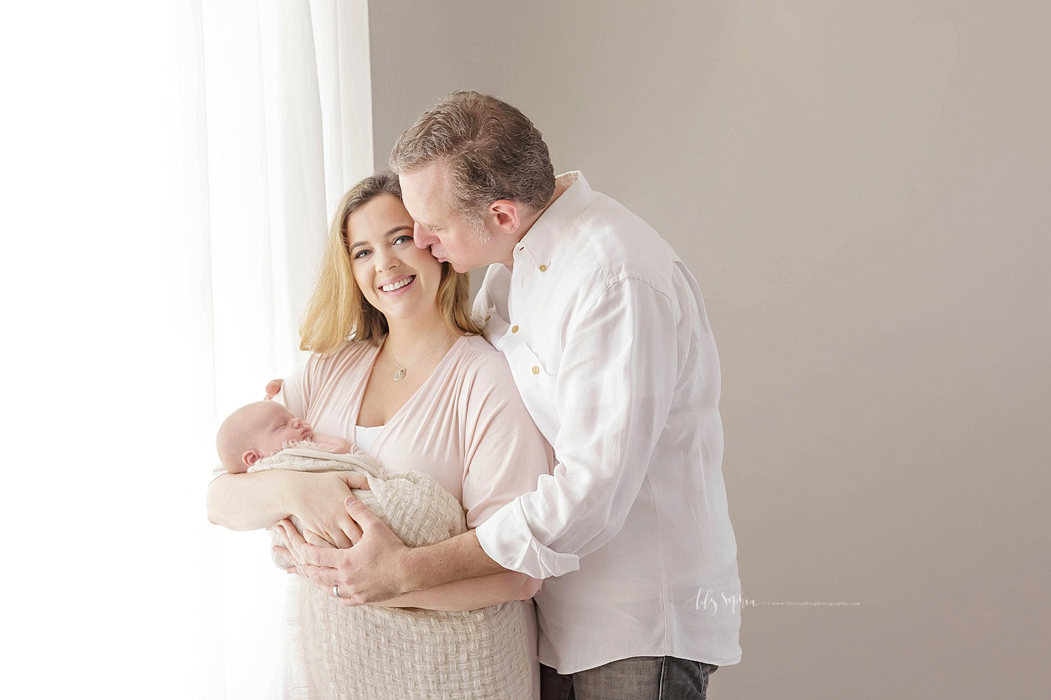  Image of a family of three. The mother is wearing a light pink dress and holding her sleeping, newborn son. The father is standing behind her, kissing her cheek, with one hand on the baby, and the other hand on her shoulder. &nbsp; 