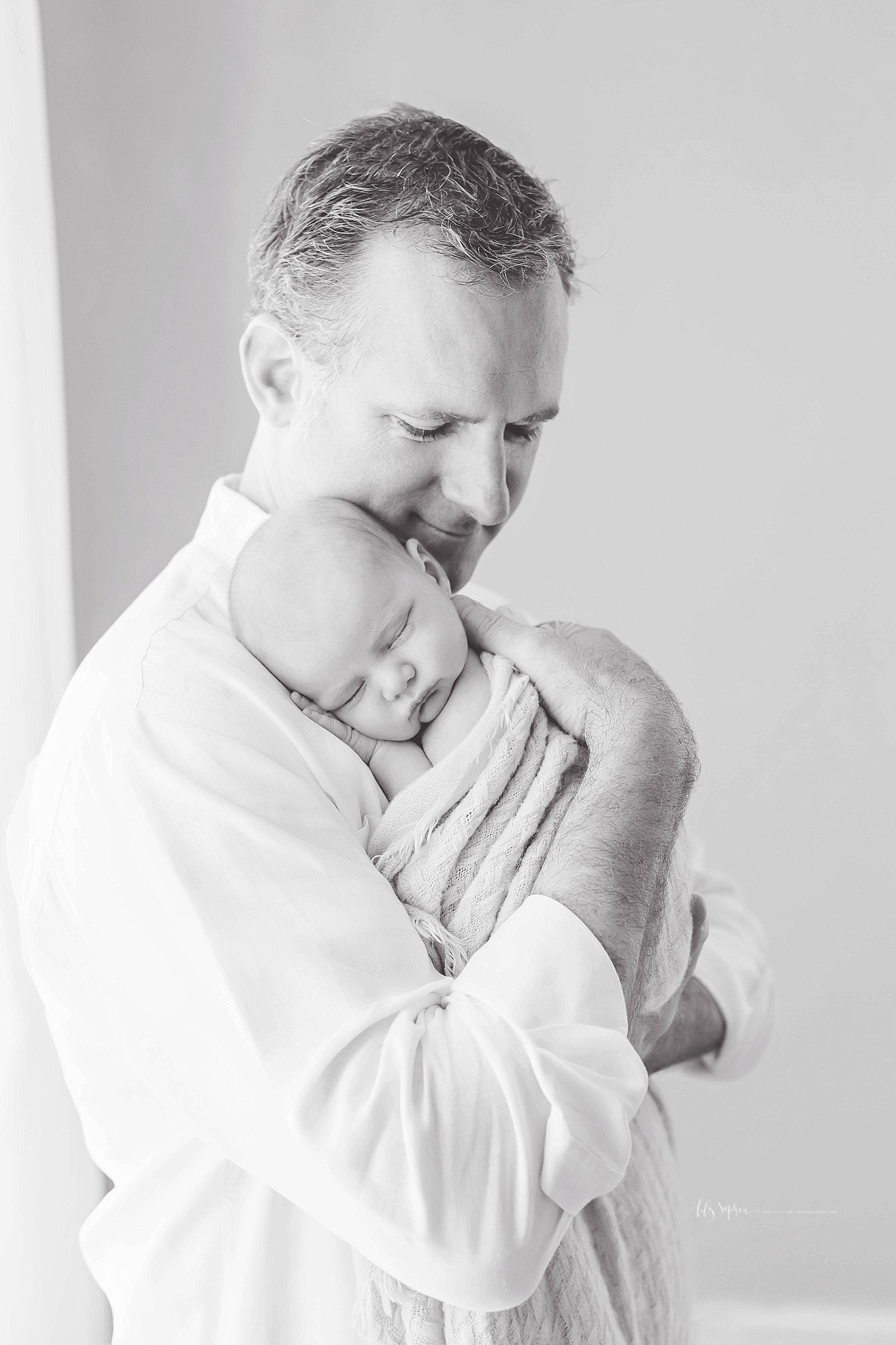  Black and white image of a father holding his sleeping, newborn son on his shoulder, while he looks down at him and smiles.&nbsp; 