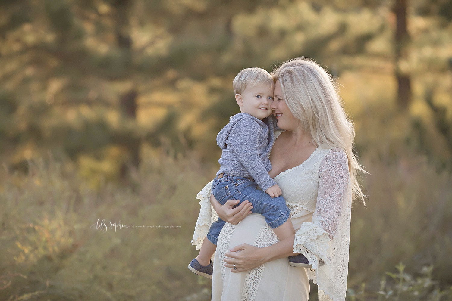  Image of a pregnant woman, in a lace, long sleeved dress, with bell sleeves, holding her toddler boy on her belly and smiling at him, while he smiles at the camera.&nbsp; 