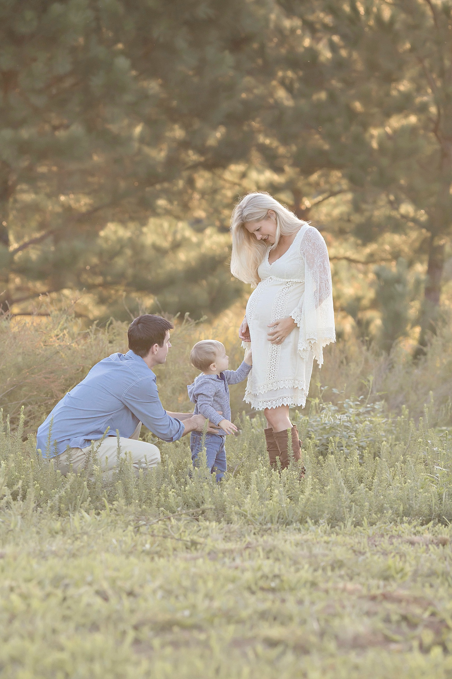  Image of a father squatting down, in a field, next to his toddler son, who has his hand on his mother's pregnant belly.&nbsp; 