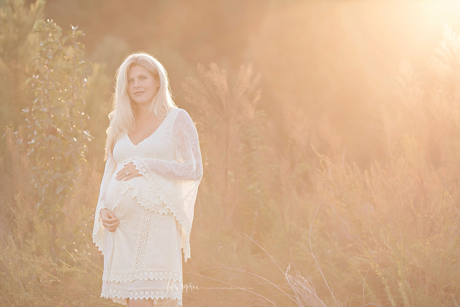  Image of a pregnant, woman, wearing a lace dress, with bell sleeves, standing in a field at sunset.&nbsp; 