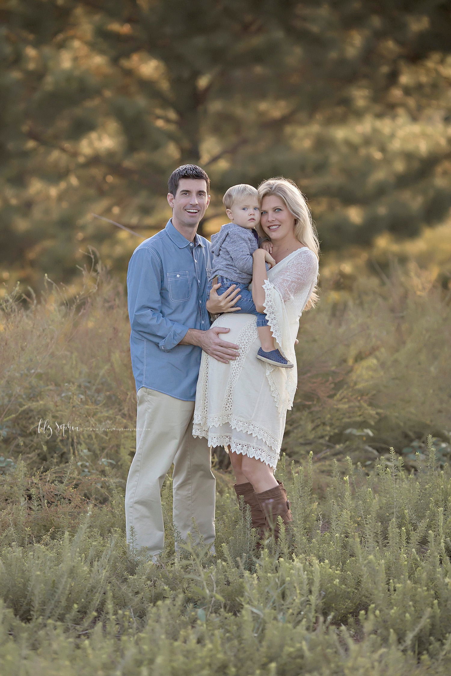  Image of a man and a pregnant woman standing in a field with their toddler son in between them.&nbsp; 