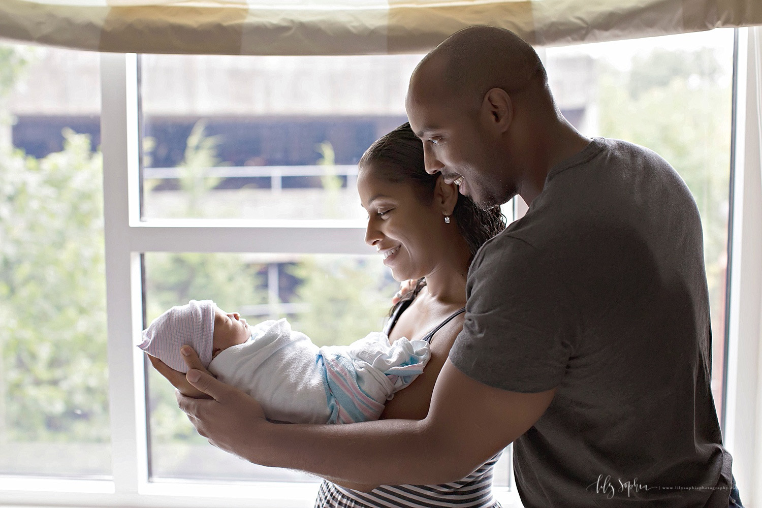  Image of African American parents, &nbsp;holding their sleeping newborn son in their arms in front of a window.&nbsp; 