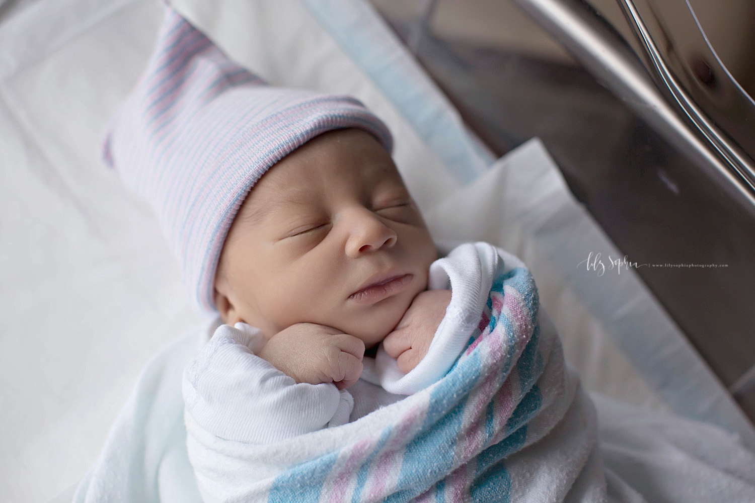  Image of a sleeping, African American, baby, boy, in his hospital bassinet, wrapped in a blanket with his hands under his chin.&nbsp; 