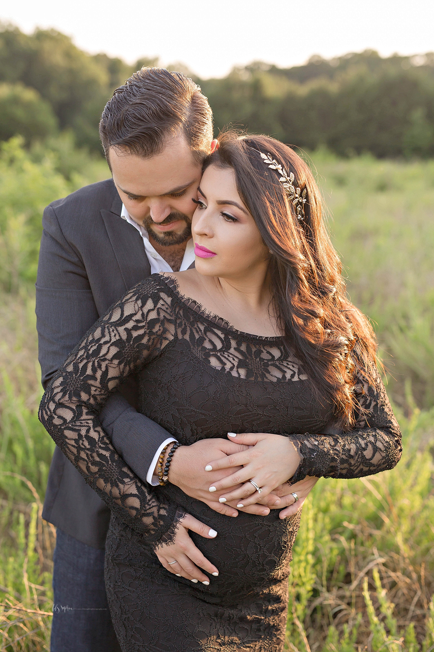  Image of a latino man, hugging his pregnant wife from behind, standing in a field, at sunset.&nbsp; 