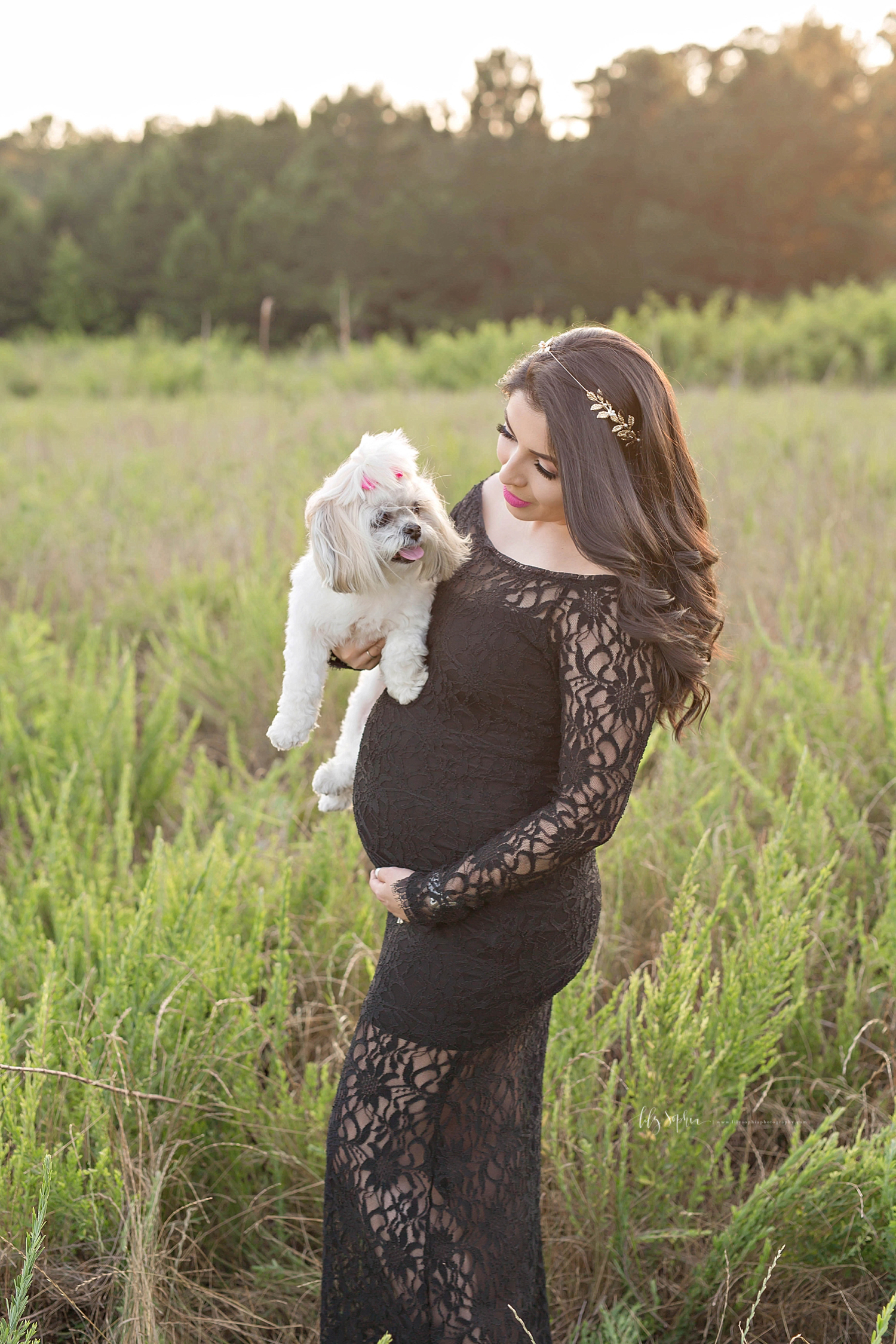  Image of a pregnant woman, in a black lace dress, standing in a field, holding her little white dog.&nbsp; 