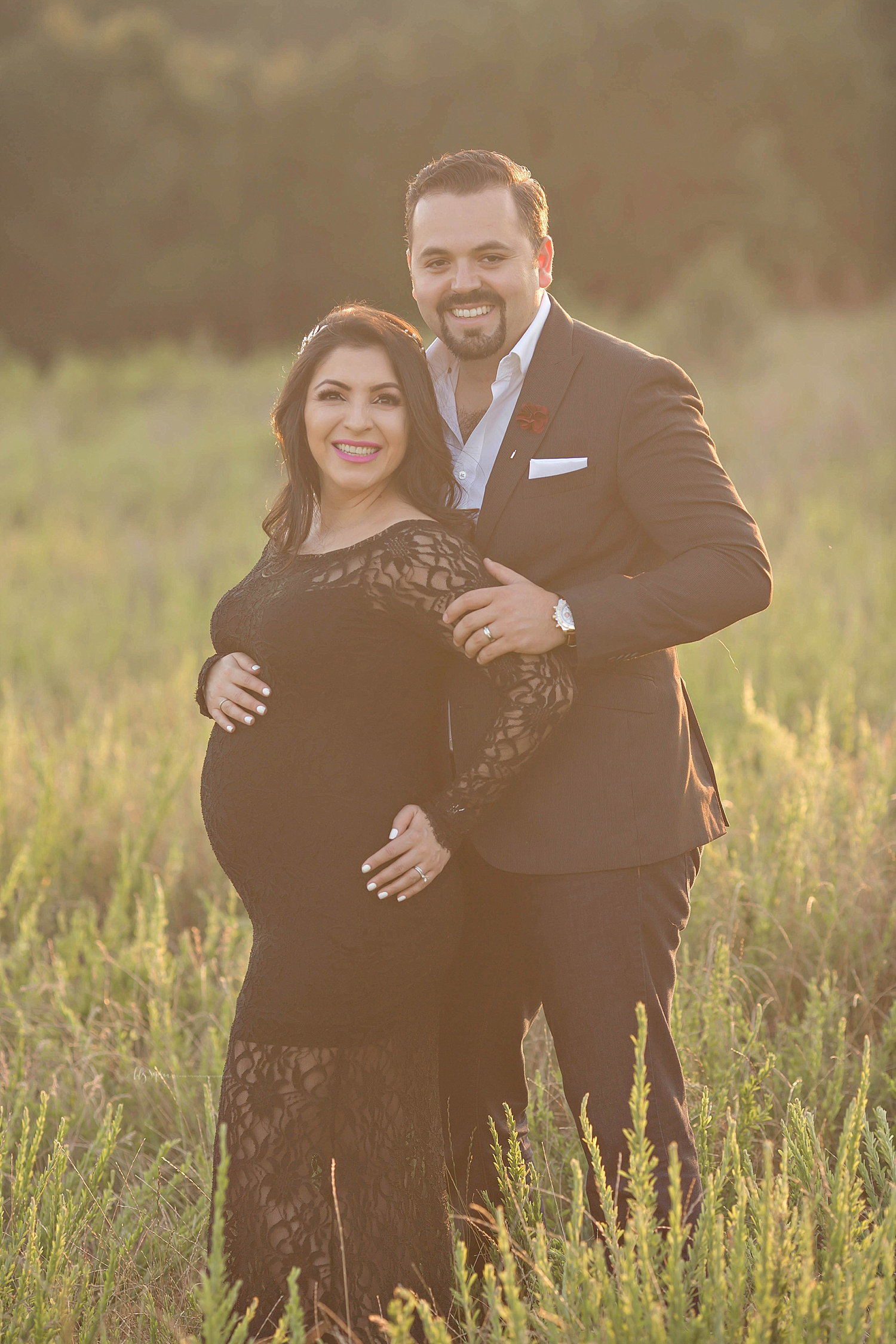  Image of a pregnant Latino couple, standing in a field at sunset, smiling.&nbsp; 