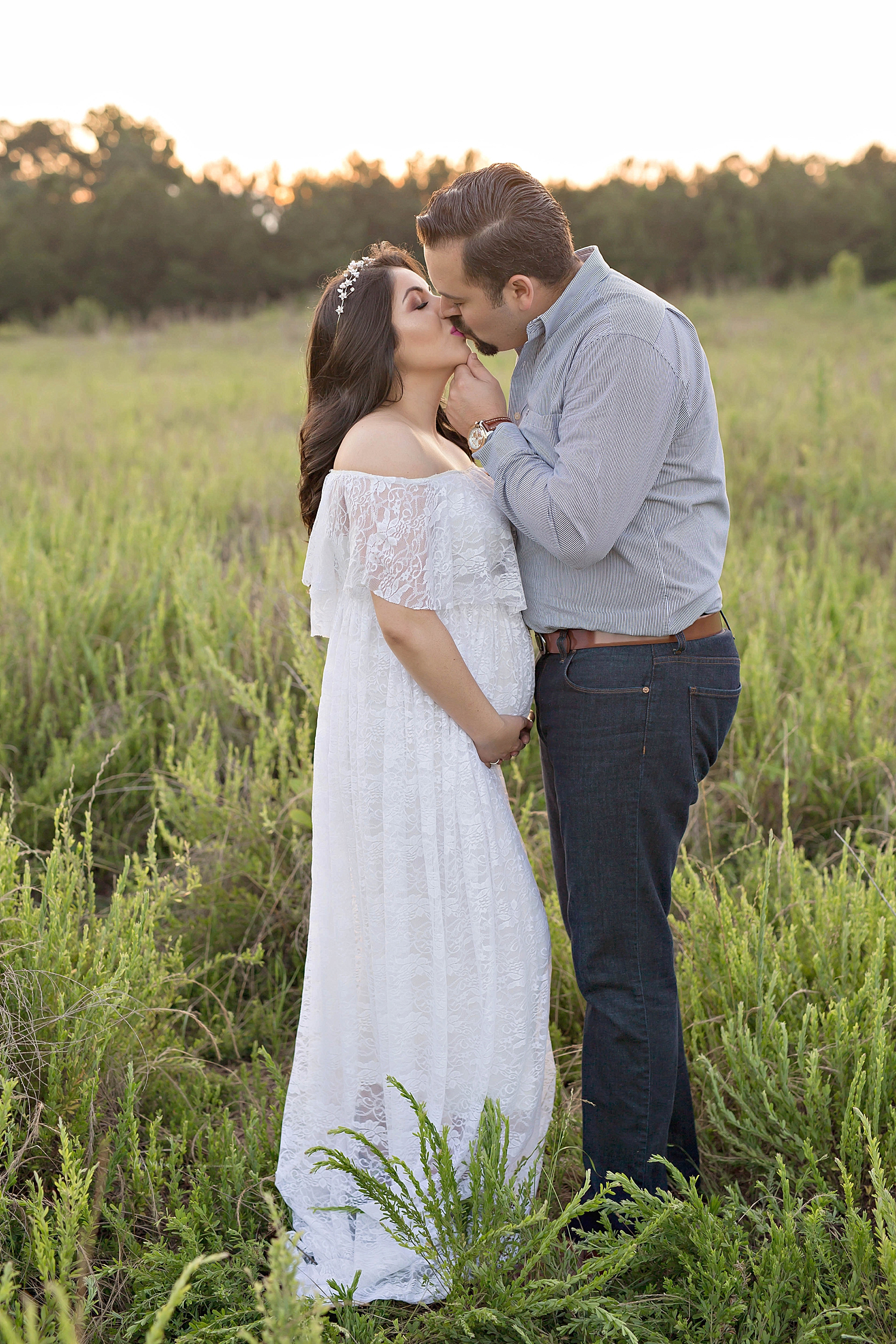 Image of a pregnant Latina woman, wearing a white, lace, off the shoulder, dress, kissing her husband in a field.&nbsp; 