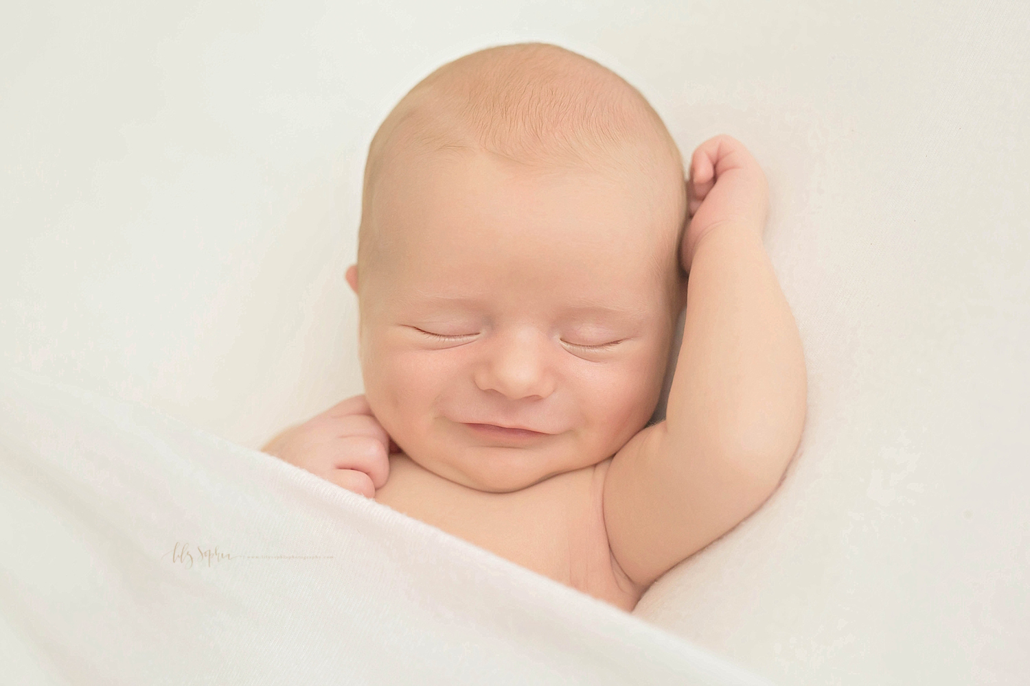  Newborn baby boy with red hair grinning as he sleeps on his back with his right hand reaching above his head.  He is lying between a white blanket. 