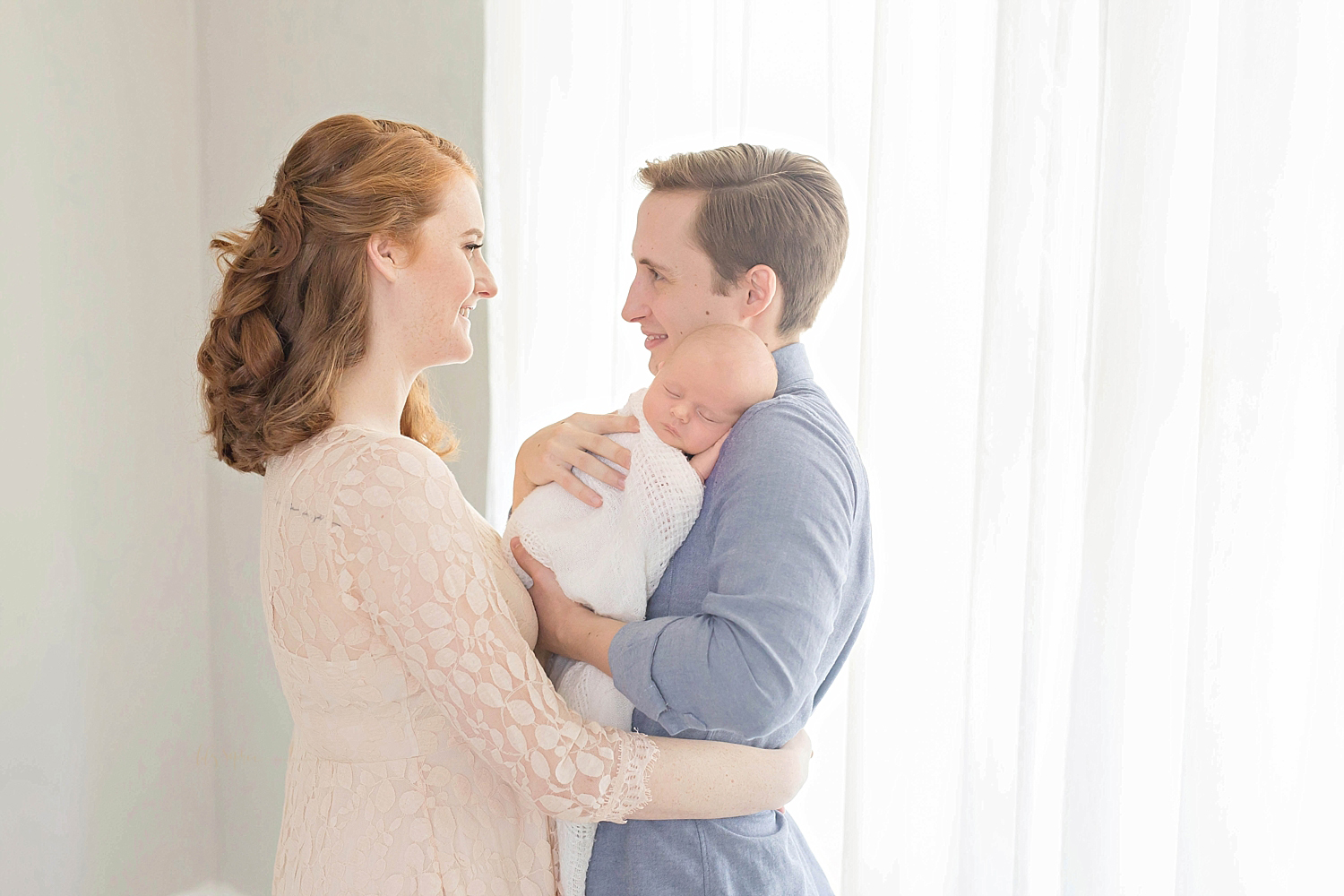  Red-haired woman wearing a blush pink lace dress stands facing her husband who is wearing a blue chambray buttoned-down shirt.  The husband holds his sleeping infant son in his arms.  The wife is holding her husband around his waist and the two of t