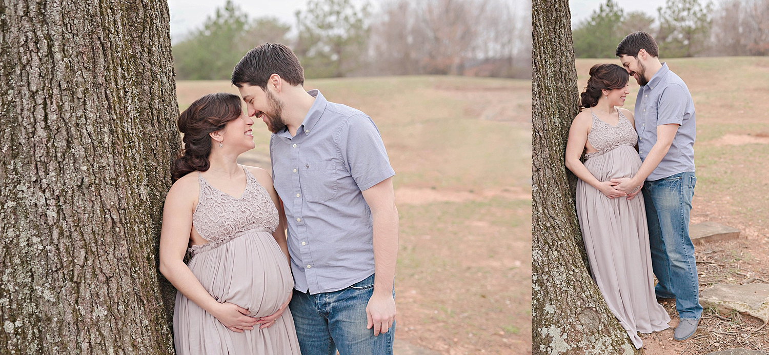  Photo of an expectant couple in a park.  The woman is wearing a taupe backless halter dress with a lace bodice.  The man is wearing a short sleeve light blue buttoned down shirt and blue jeans.  The woman is standing against a tree as her husband lo