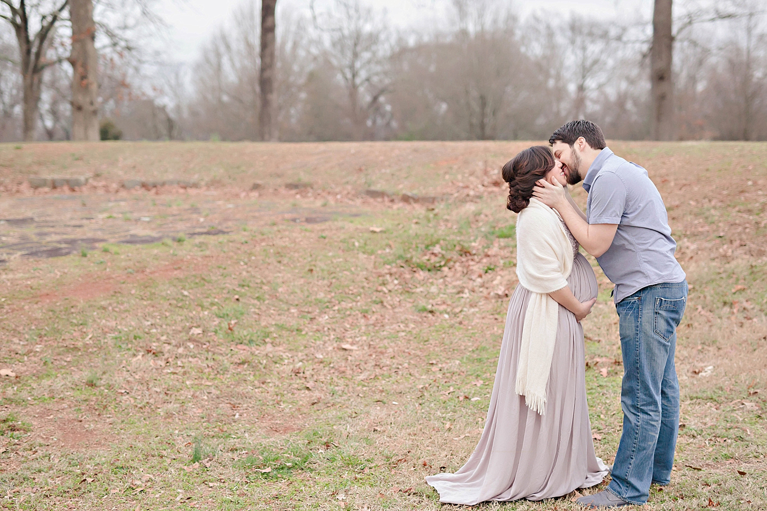  The expectant couple are standing in an Atlanta park.  The husband holds his wife’s head in her hand as he kisses her. 