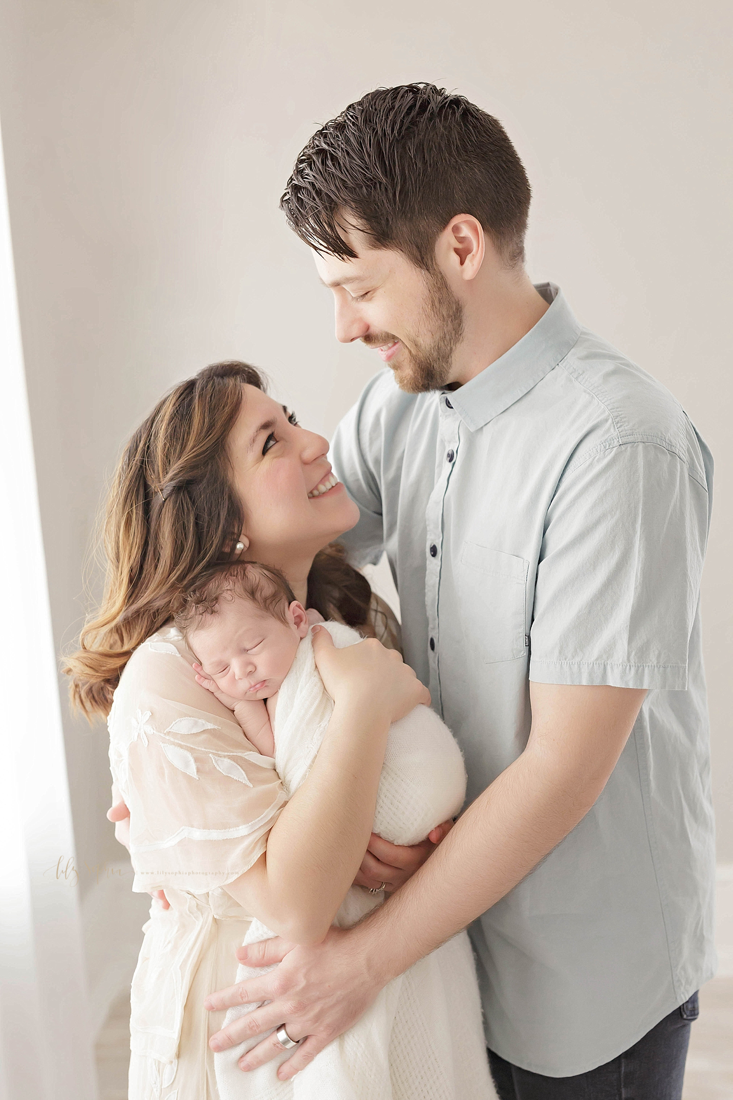  Mom and Dad look lovingly at one another in an Atlanta studio.   Mom has long auburn hair and is wearing a blush pink flowing dress.  Her bearded husband who is wearing a buttoned-down short sleeve blue shirt  looks down at her as she holds their in