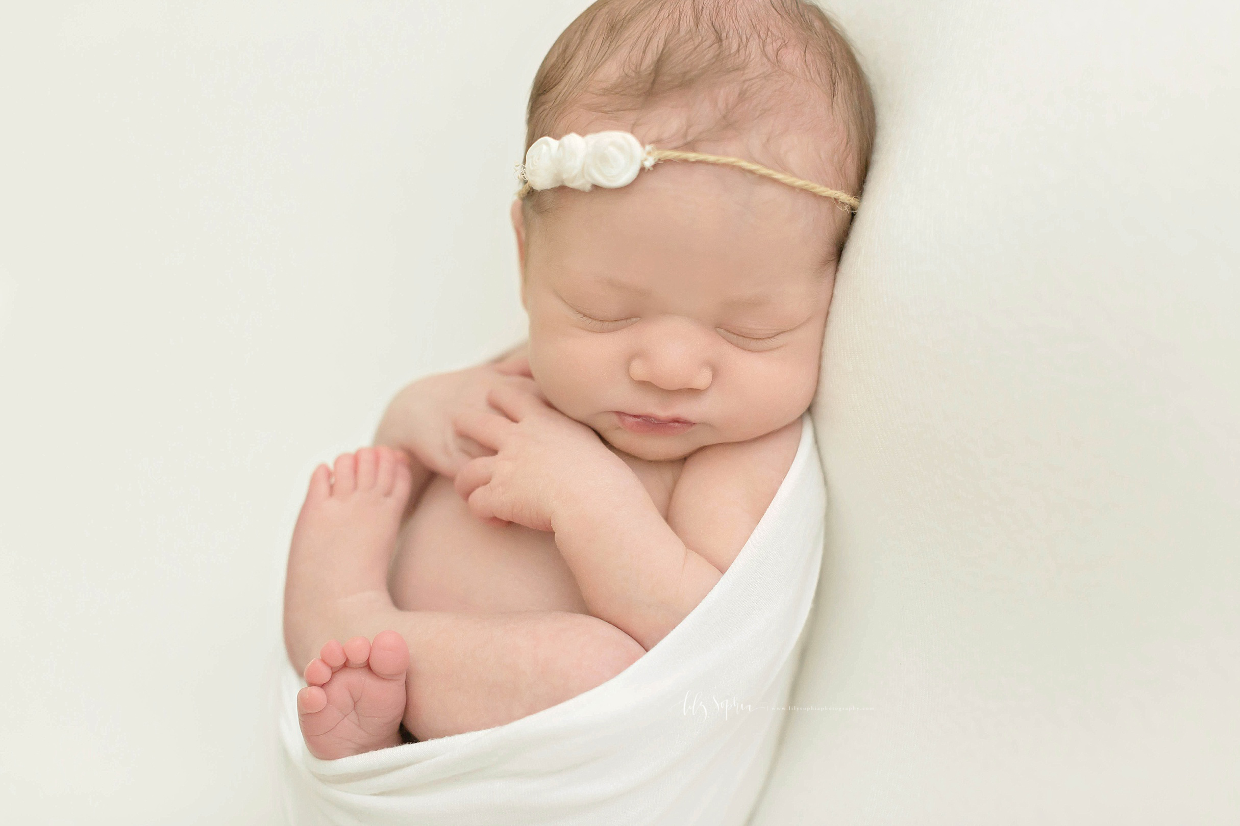  Image of a sleeping, newborn, baby, girl, with a delicate rosebud tie back in her hair, wrapped in the egg pose.&nbsp; 
