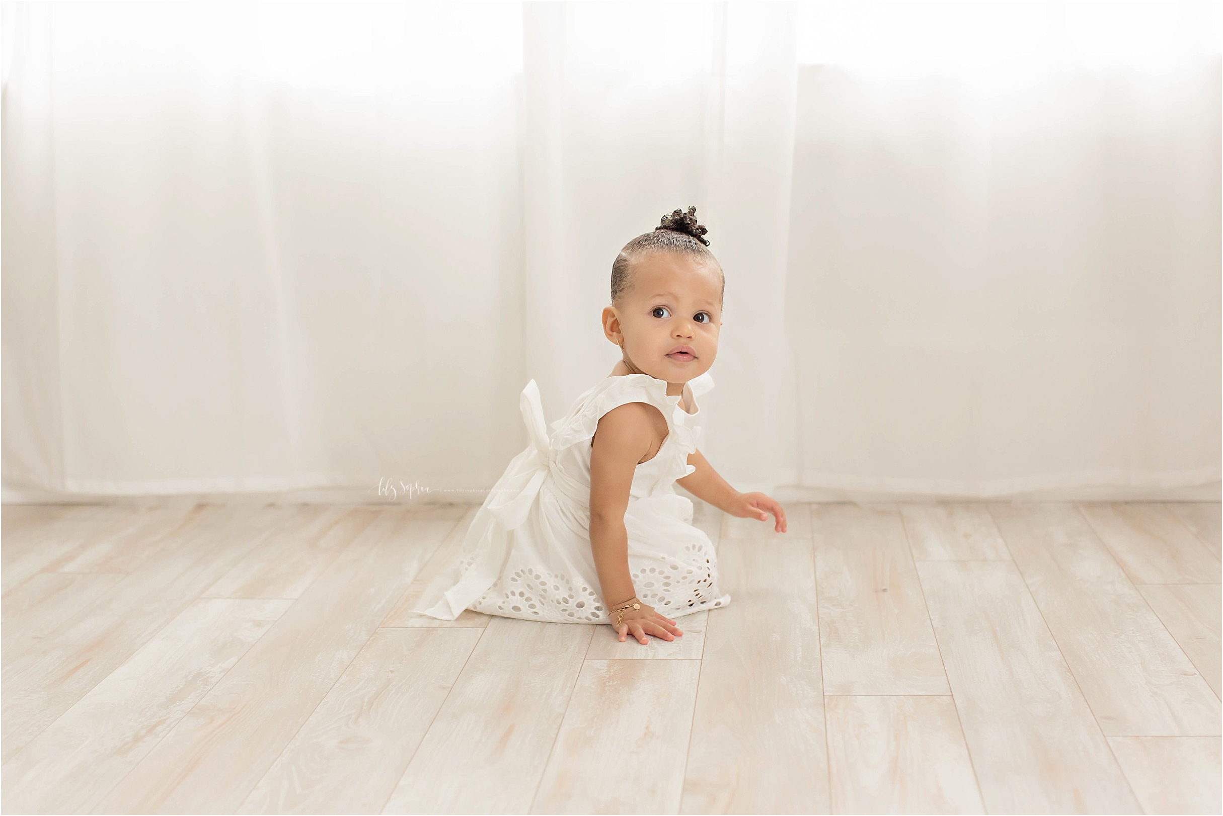  Image of an African American, baby, girl, wearing a white eyelet dress, sitting on the floor.&nbsp; 