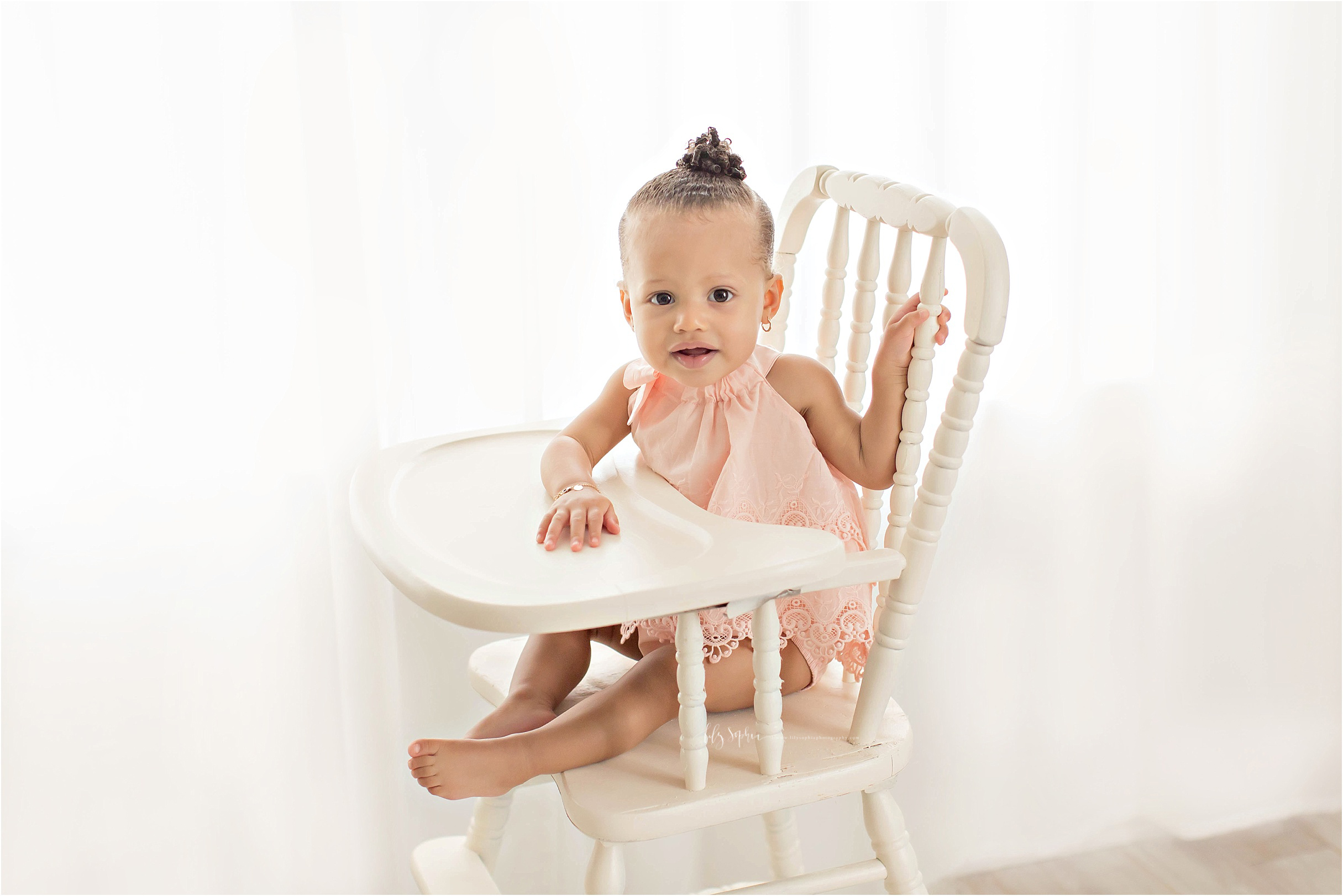  Image of an African American baby girl, with a bun in her hair, wearing a peach dress and sitting in an antique wooden highchair.&nbsp; 