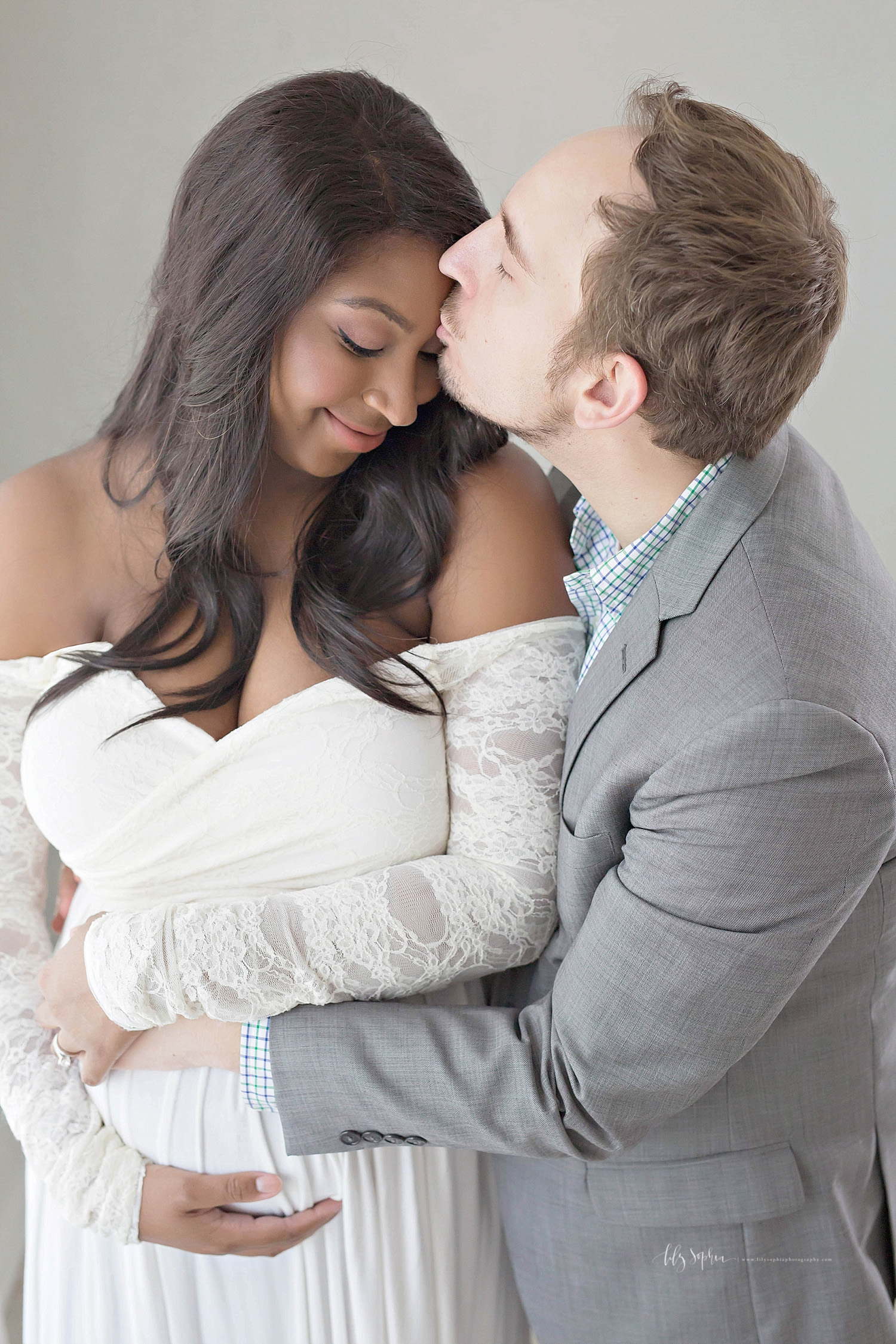  Image of an African American, pregnant, woman, wearing an off the shoulder, cream dress with lace sleeves, looking down and smiling, while her Caucasain husband kisses her forehead.&nbsp; 