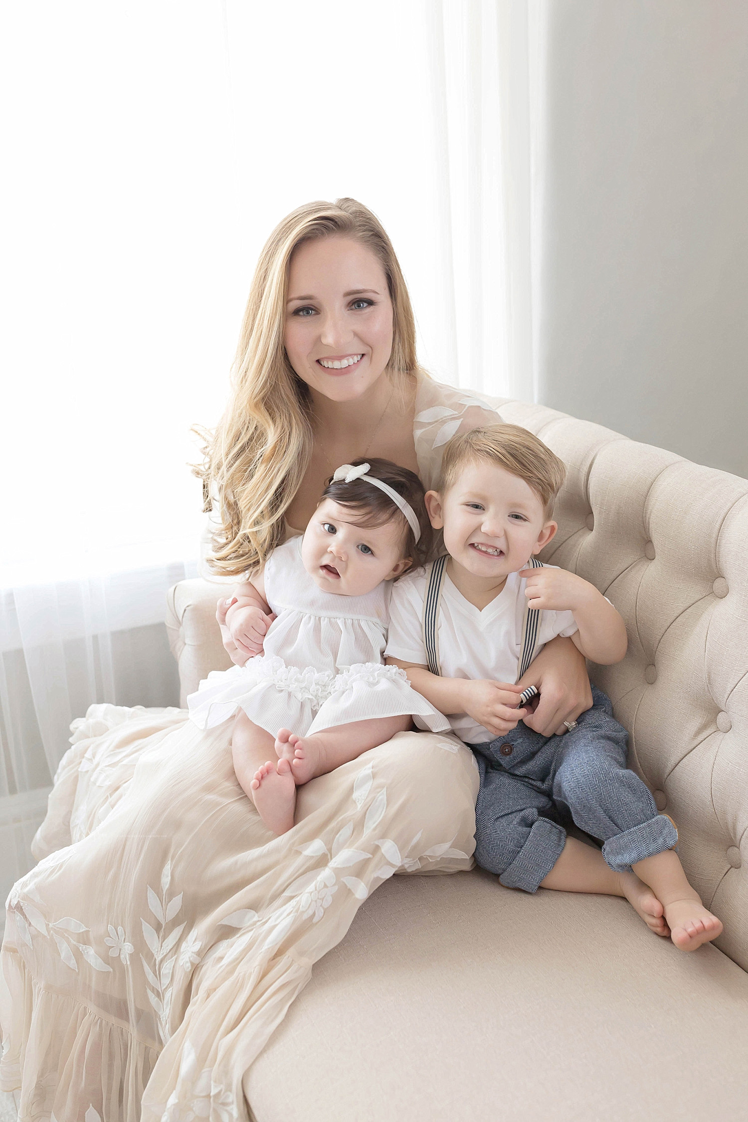  Image of a mother, smiling at the camera, while holding her baby girl and toddler son on her lap. 