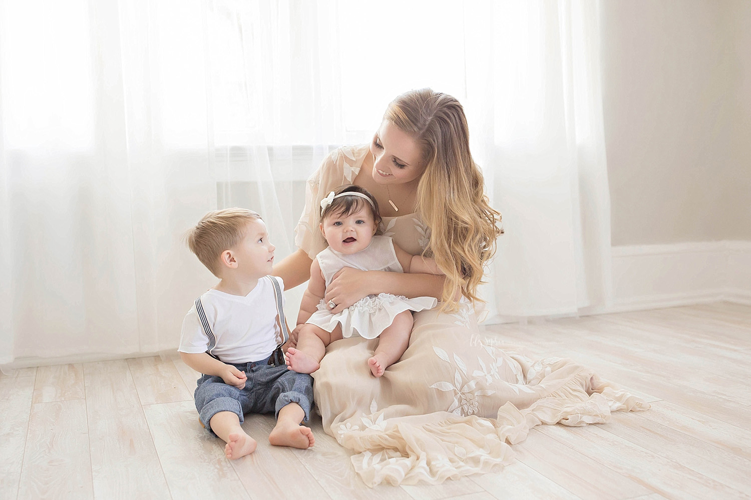  Image of a mother, sitting on the floor in a taupe dress, with her daughter on her lap, and her son next to her.&nbsp; 