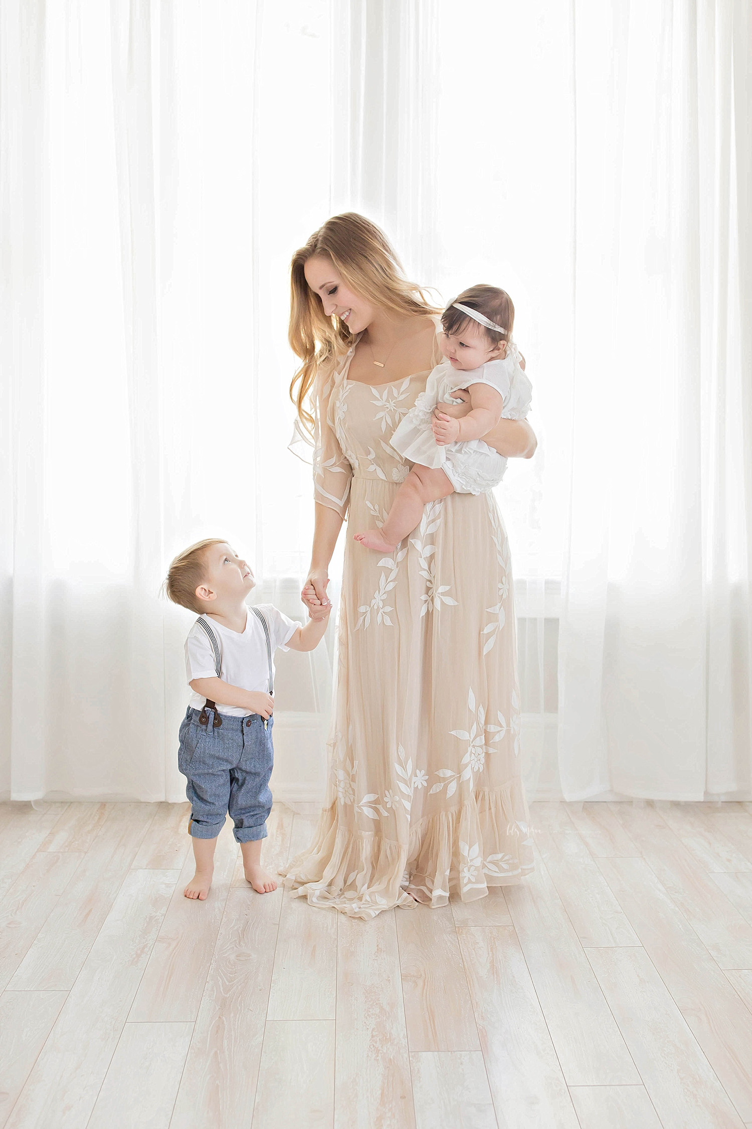  Image of a mom, holding her son's hand while they look at each other and she balances her daughter on her hip. 