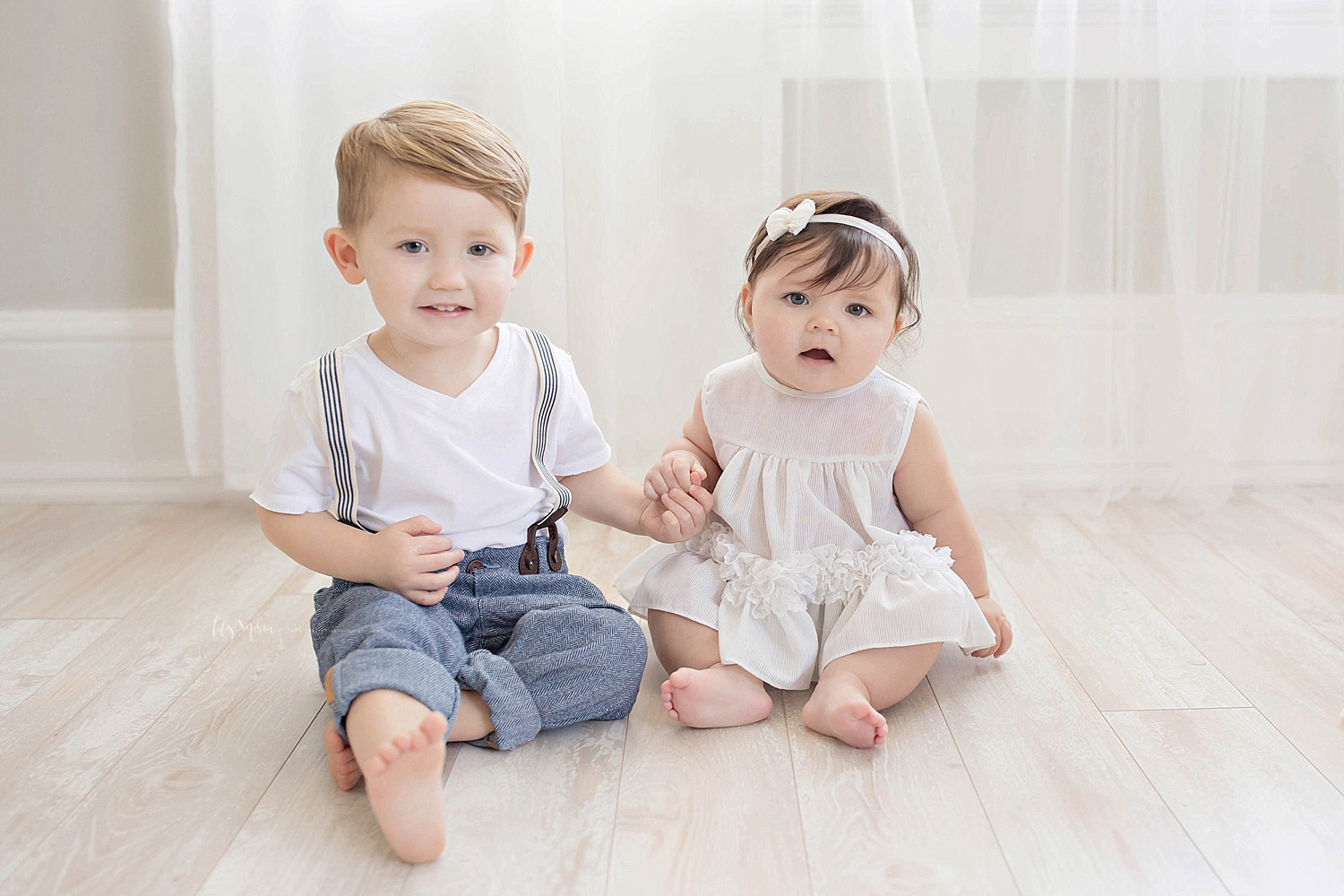  Image of a toddler boy in rolled up jeans and suspenders, sitting on the floor and holding his baby sister's hand.&nbsp; 