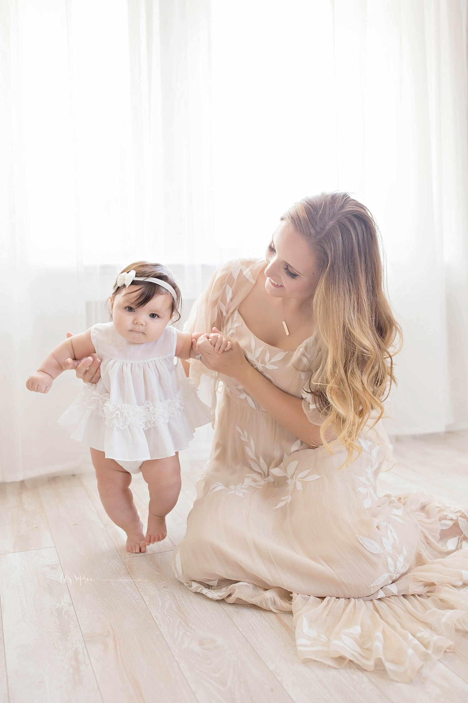  Image of a mother, wearing a taupe dress, helping her baby girl stand up.&nbsp; 