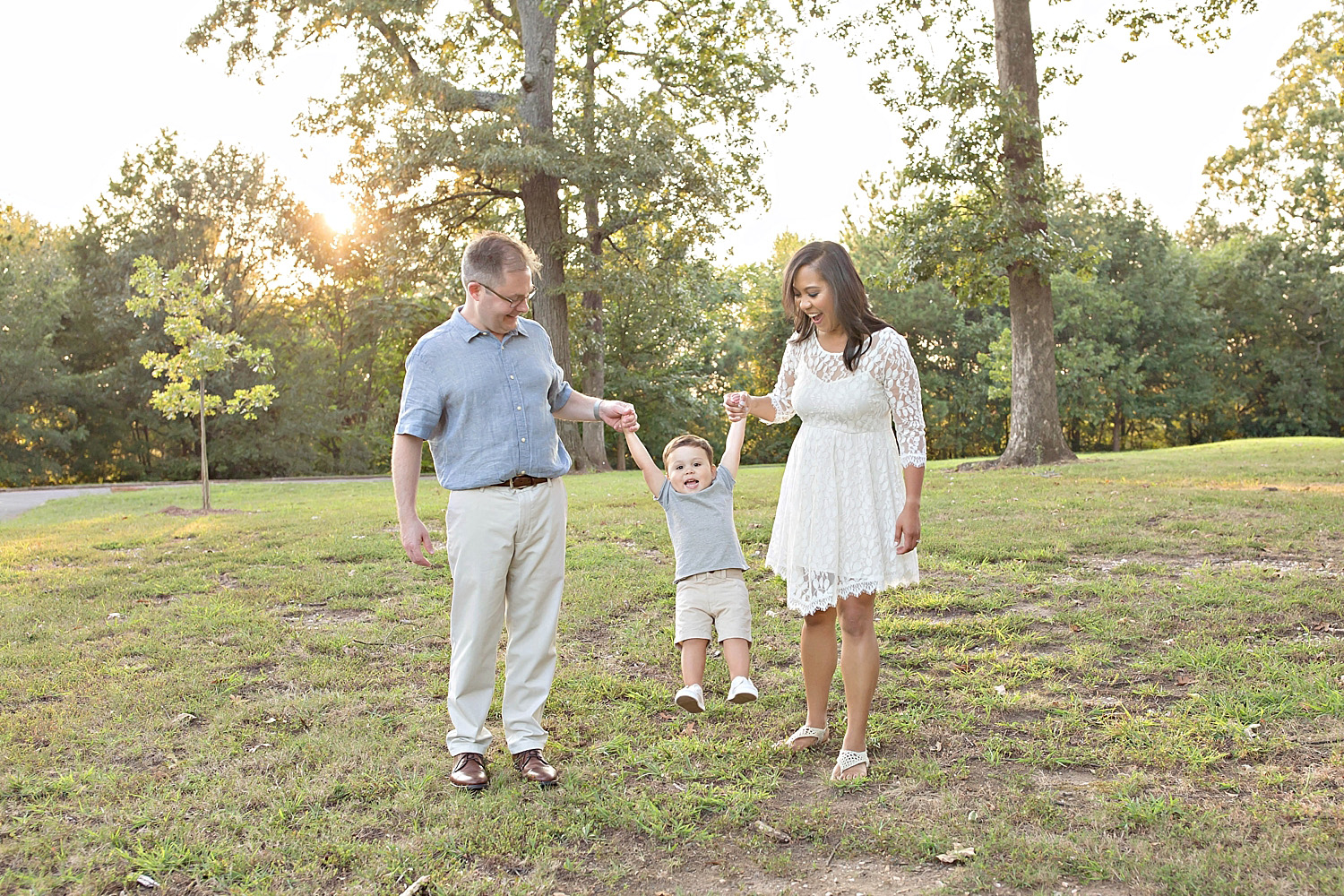  Image of a mother and father, standing in a park at sunset, with their son in between them, holding him up by his arms and swinging him.&nbsp; 