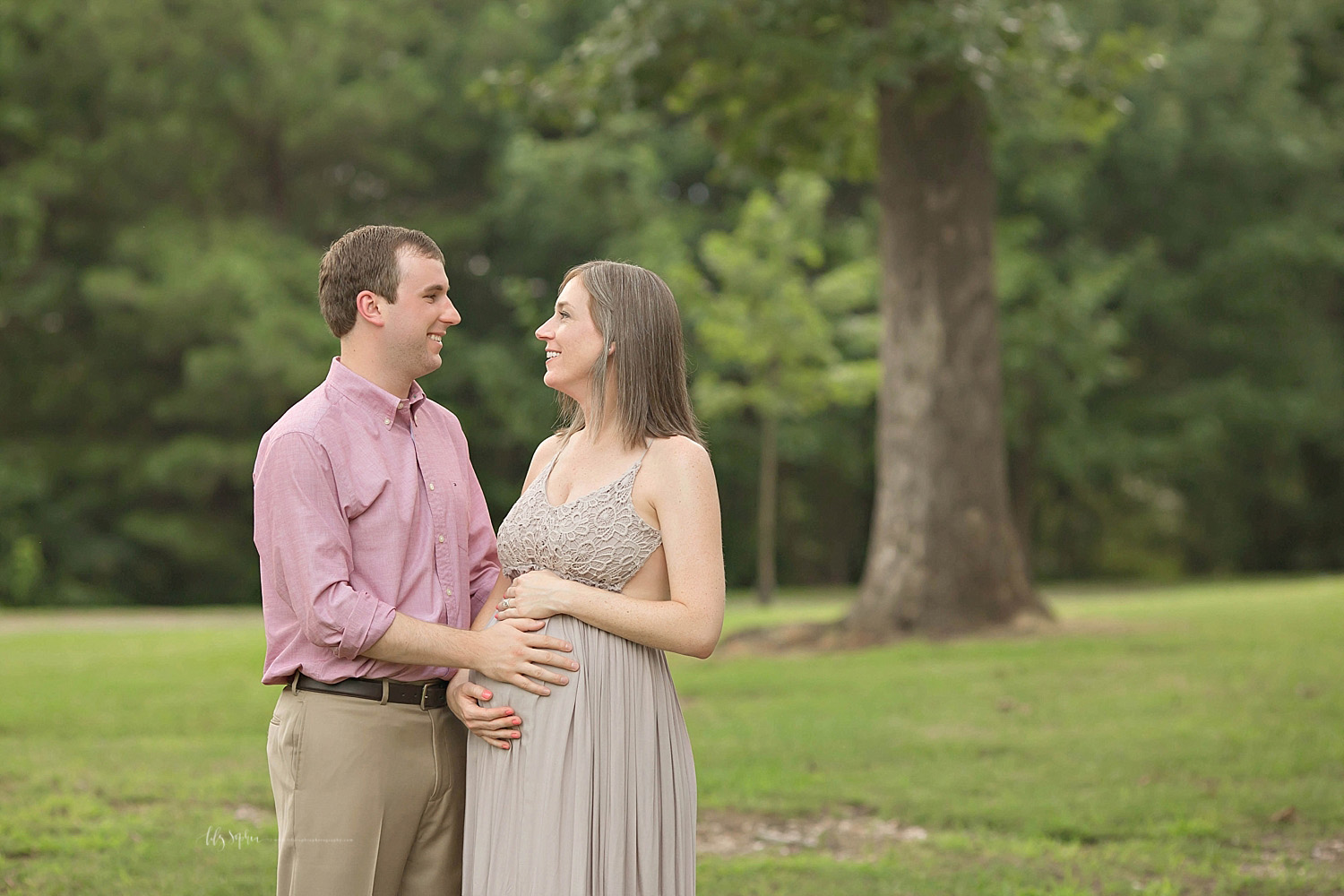  Image of a pregnant woman, holding her belly and looking at her husband and smiling at him while they stand in a park.&nbsp; 