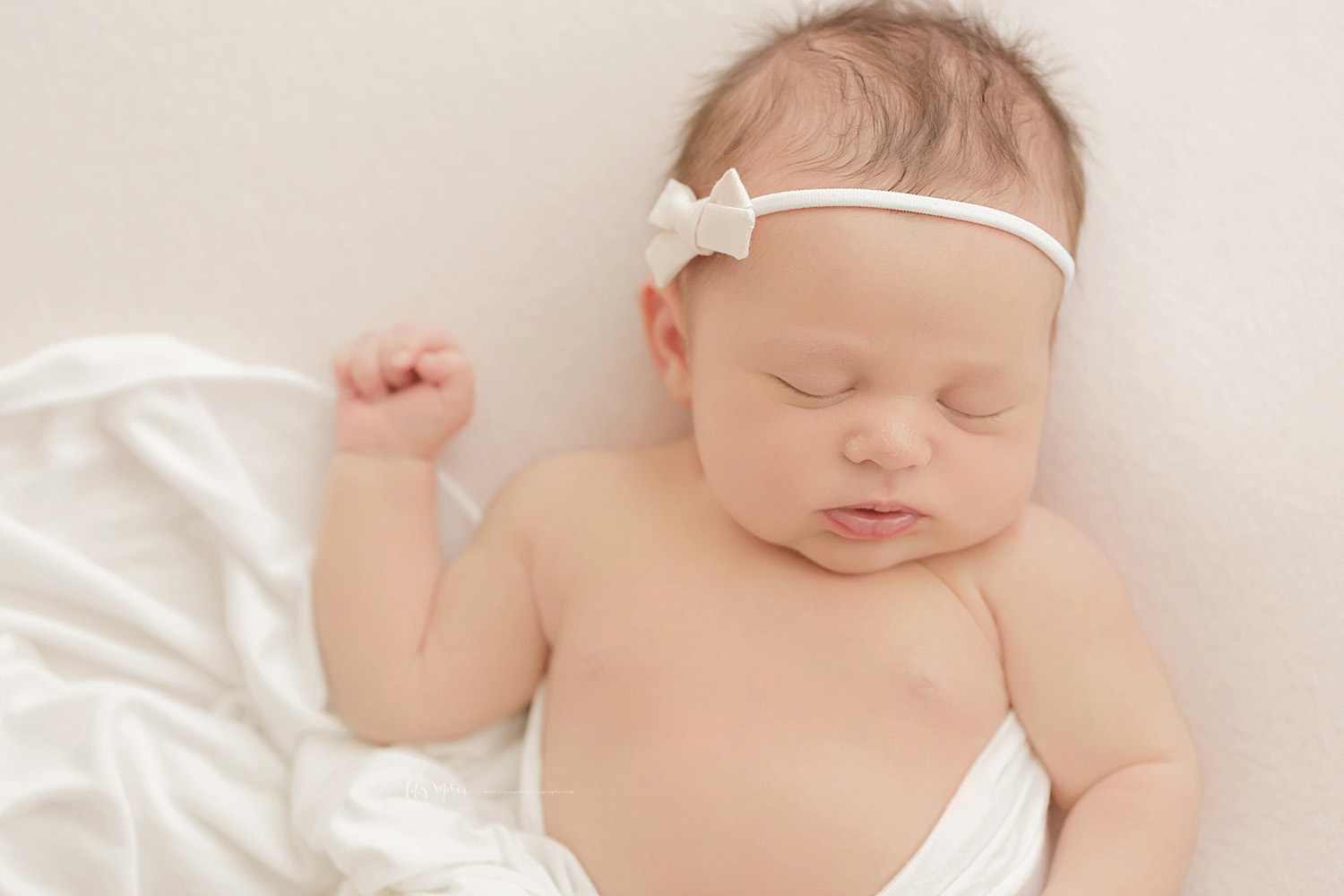  Image of a sleeping, newborn, baby, girl, with a white bow headband on her head, and lay on her back with one arm up by her head.&nbsp; 